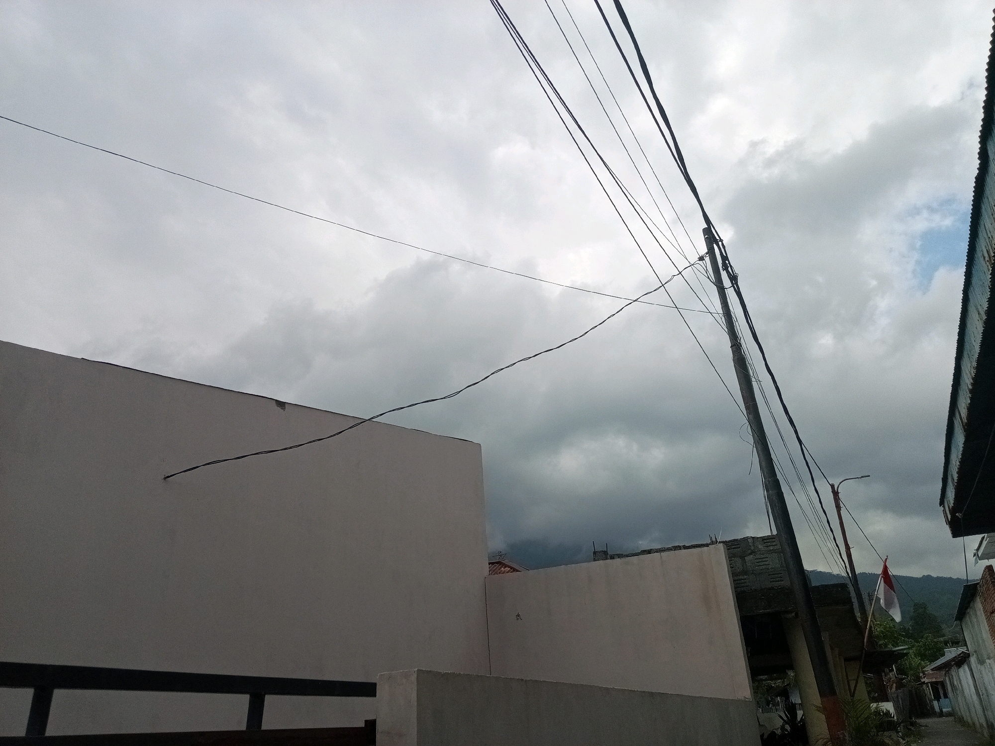 The image depicts a cloudy sky over a residential area in Bitung, Indonesia. In the foreground, a large, light-colored building occupies the left side of the frame, with a dark railing along its lower edge. To the right of this building is another structure of similar color. A utility pole with numerous wires stretches diagonally across the sky, originating from the right side of the image and extending towards the upper left. A small Indonesian flag, red and white, is attached to the utility pole. In the background, there are more houses, some with corrugated metal roofs, and a street. Distant hills are visible beneath the overcast sky. The overall atmosphere is subdued due to the cloudy weather.