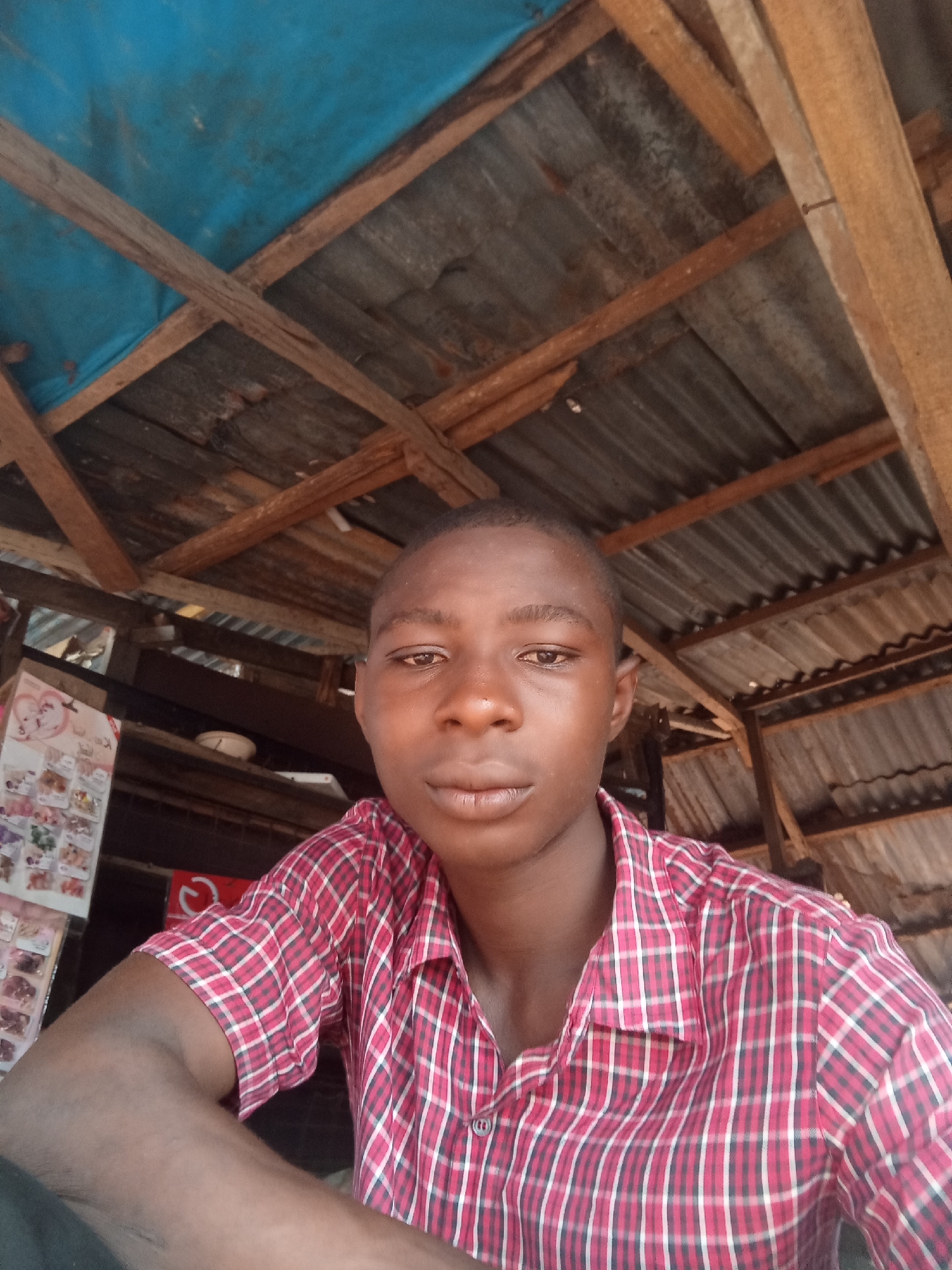The media file shows a young person, likely a male, in a close-up, low-angle selfie. The subject is wearing a red and white plaid shirt. Their gaze is directed towards the camera, and their expression appears neutral.
The setting suggests an outdoor or semi-enclosed space with a rustic roof structure. Wooden beams and corrugated metal sheeting are visible, forming a makeshift ceiling. A blue tarp is also present overhead, adding to the impression of an informal construction.
In the background, to the left of the subject, there is a display of various items, possibly small products or trinkets, arranged on shelves or a board. Some of these items appear to be packaged or in small containers, with what looks like printed designs on them. A red circular object with a white symbol is also visible behind the subject's arm.
The lighting suggests it is daytime, though the ambient light is somewhat dim, possibly indicating an overcast sky or a shaded area. No specific weather conditions can be inferred. The overall impression is a candid snapshot taken in a simple, possibly market or roadside stall environment. There is no visible text in the image that is clear enough to be read.