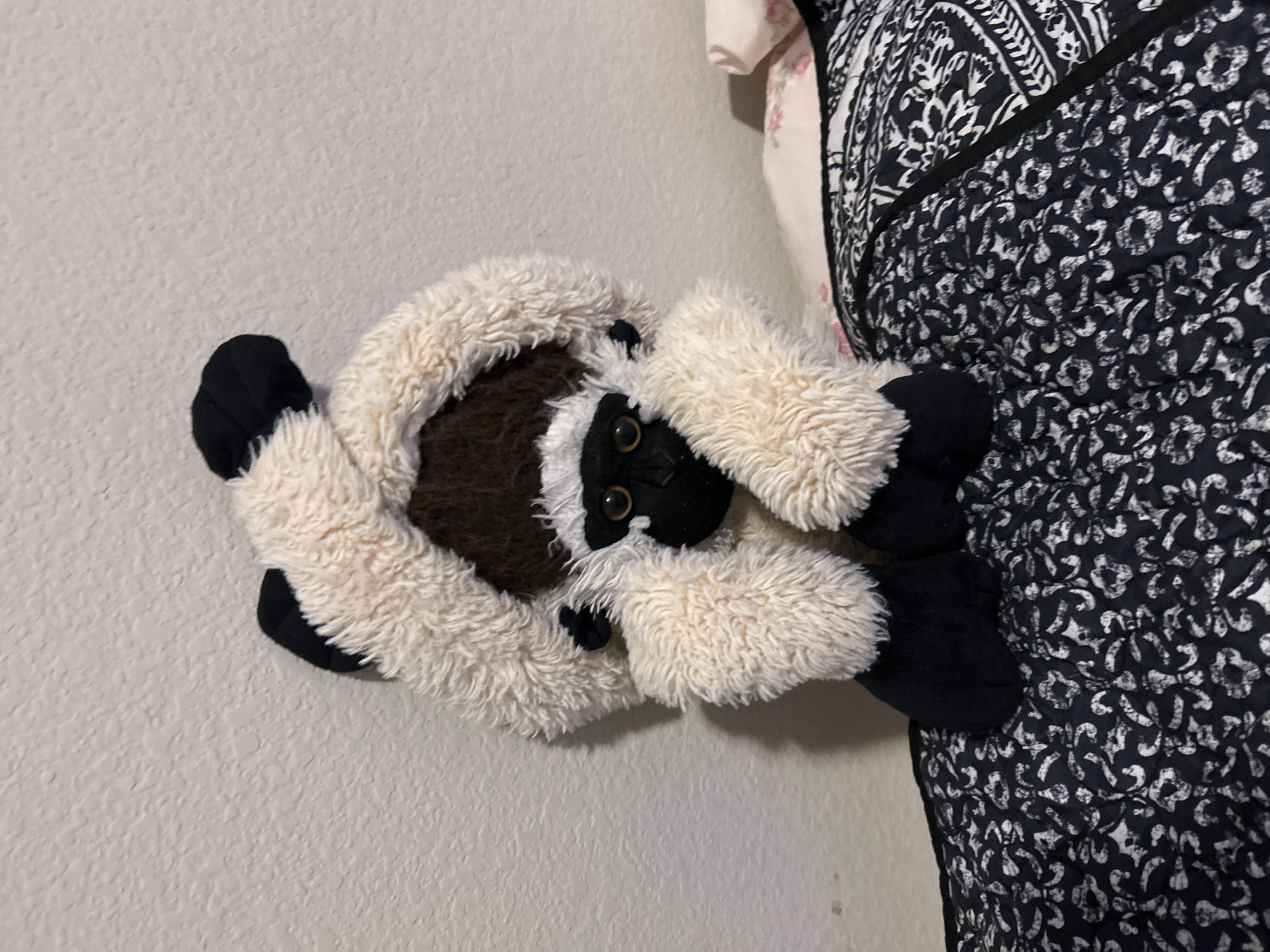 A stuffed primate with cream-colored fur and black accents is centered on a black and white patterned quilt. The stuffed animal has a black face, large expressive eyes, and is wearing a cream-colored, hooded costume. The background is a textured white wall. The quilt is on a bed, and a portion of a pink floral pillow is visible to the right of the stuffed animal. The scene appears to be indoors, likely in a bedroom.