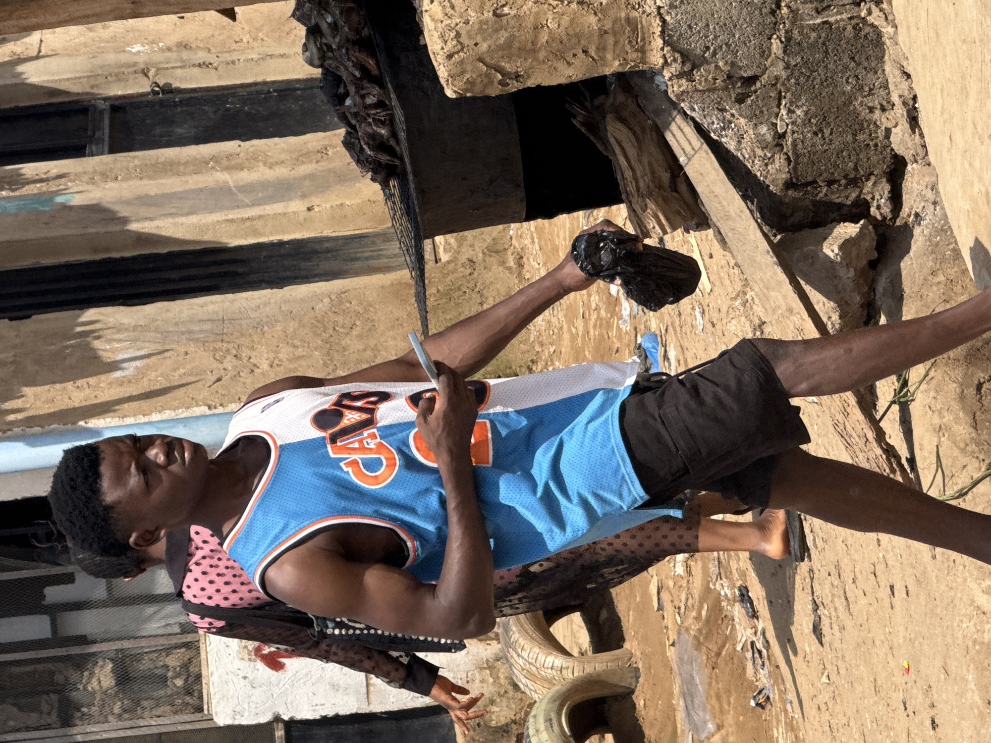 The image depicts a street scene in Abuja, Nigeria, with a young Black male in the foreground. He is wearing a blue and white basketball jersey and black shorts, holding a black plastic bag and a cell phone in his right hand. Behind him, a woman in a pink polka-dot dress is walking away. The background shows a rustic structure with a metal grill containing cooked meat, suggesting a food vendor or preparation area. The ground is dirt and debris, indicative of an outdoor market or informal settlement. The overall atmosphere appears to be that of a busy, everyday street in a Nigerian city.