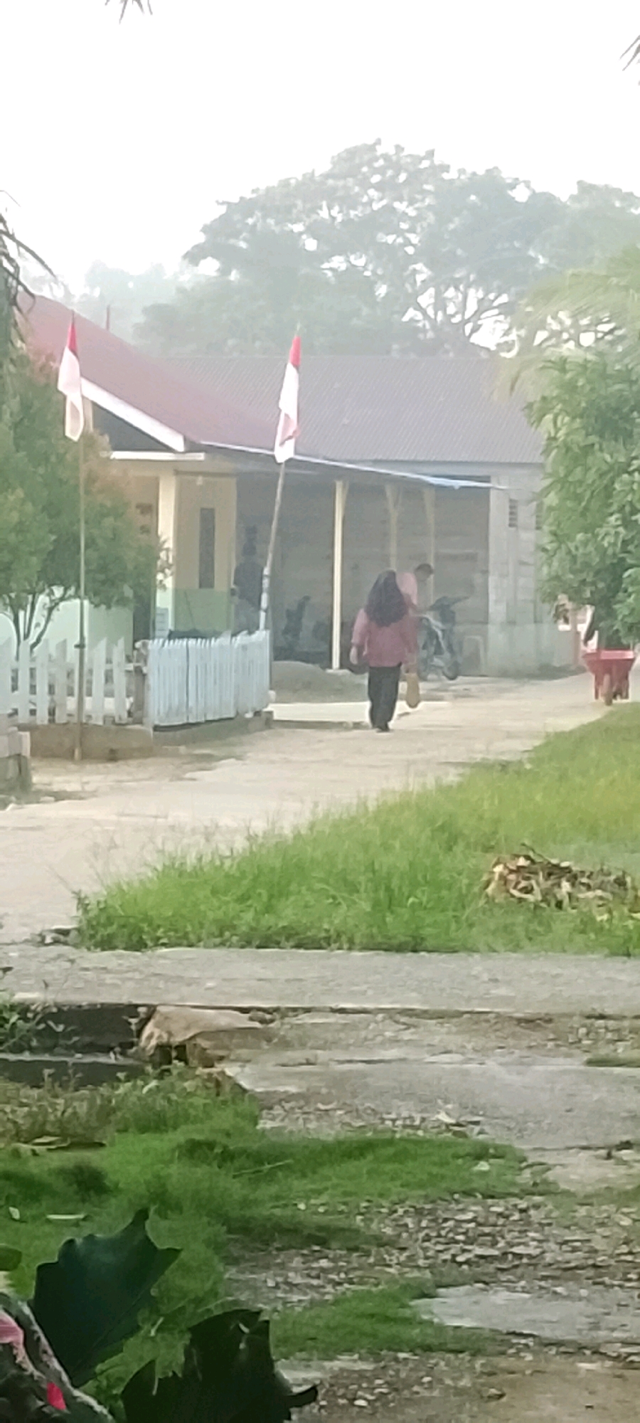In the city of Harapan Jaya, Indonesia, a scene unfolds in front of a low-rise building with a reddish-brown roof, adorned with two flags displaying red and white bands. A white picket fence demarcates the edge of the property. A person with dark hair and a light pink top walks away from the camera, followed by another person in a pink shirt. A motorcycle is parked near the building, and a red wheelbarrow is visible to the right. The ground is a mix of dirt and pavement, with patches of grass and debris. The environment appears somewhat hazy, suggesting a morning or overcast day.