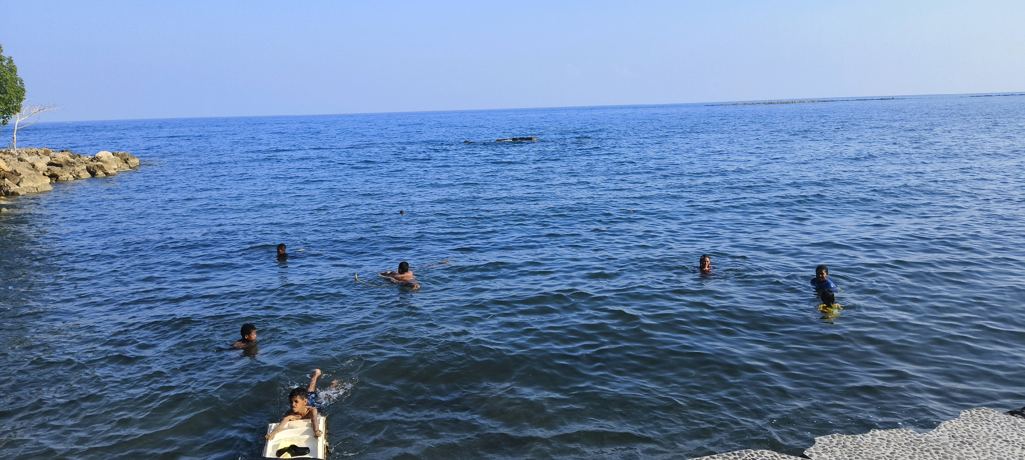 The image depicts a wide expanse of calm blue ocean under a clear sky, likely near the city of Waisai, Indonesia. Several individuals are visible swimming and interacting in the water. In the foreground, two young boys are in the water, one of whom is partially submerged in a white flotation device. To the left, a rocky shore with a single tree is visible, with a line of rocks extending into the water. Further out in the sea, at least five other individuals are swimming, with some appearing to be children. One person is floating on their back, and another is partially submerged. In the distance, a faint line of what appears to be structures or land is visible along the horizon. The water is rippled, suggesting a gentle breeze.