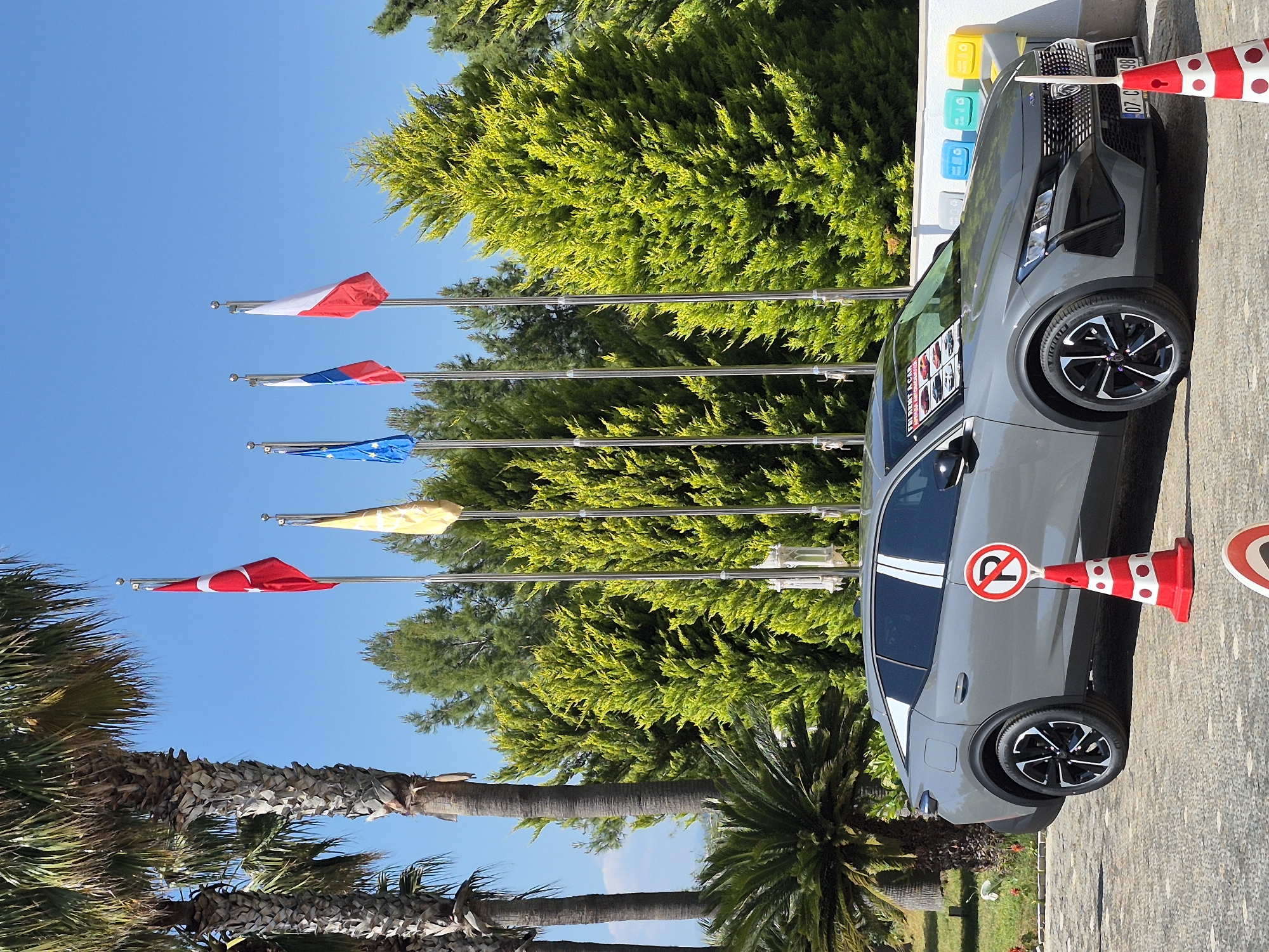 The image shows a gray car parked in what appears to be a commercial area in Alanya, Turkey, given the presence of the Turkish flag among other international flags. The car has a "Rent a Car" sign visible in the window and is flanked by traffic cones with "no parking" symbols. Behind the car are recycling bins. The area features palm trees and other greenery under a bright blue sky.
