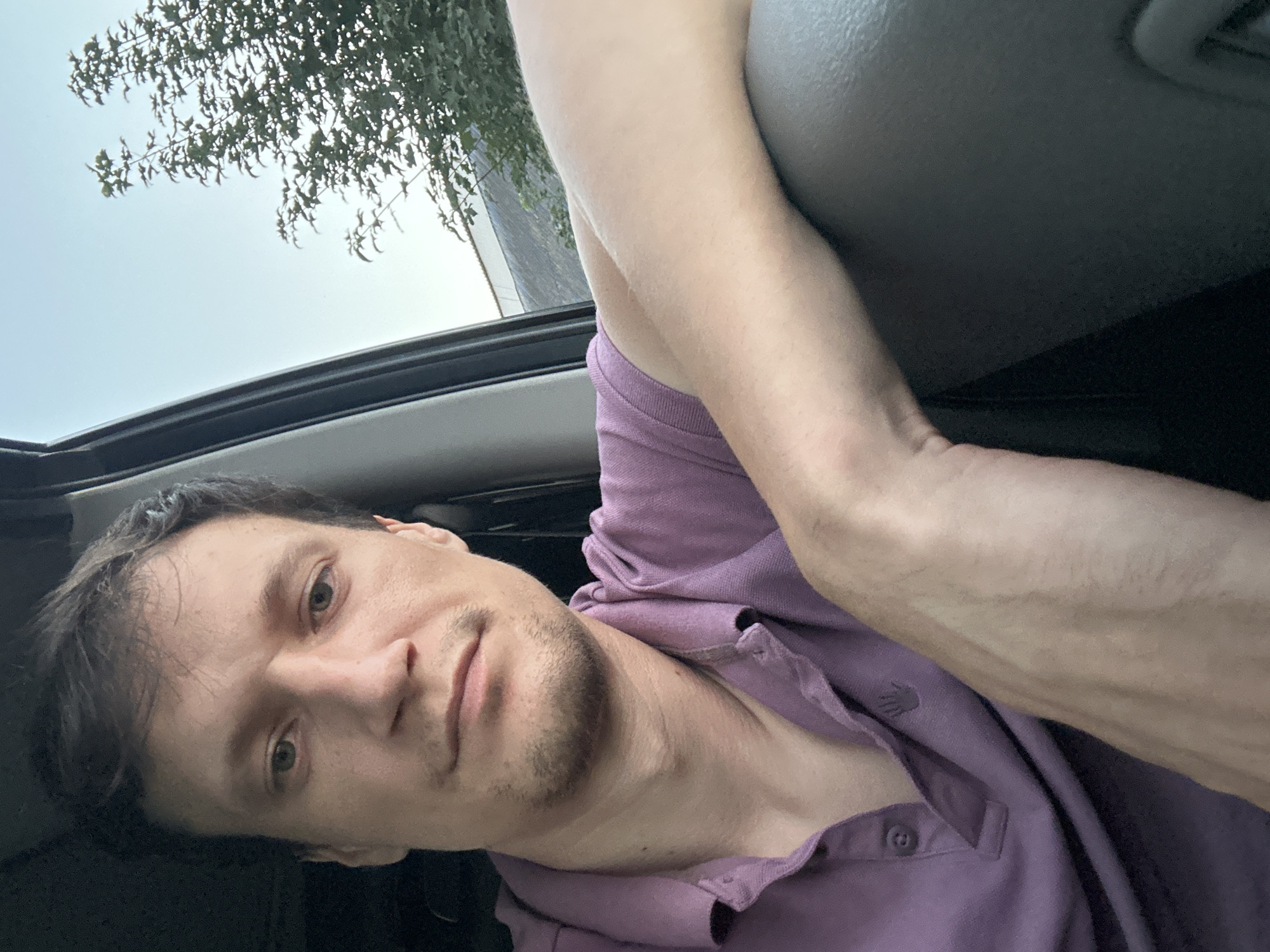 A man is captured in a selfie-style portrait from the chest up, seated inside a vehicle. He is wearing a purple polo shirt. His left arm is resting on the car's interior. The background shows the sky and greenery, suggesting an outdoor setting. The image does not contain any information about the location in Shatava, Ukraine.