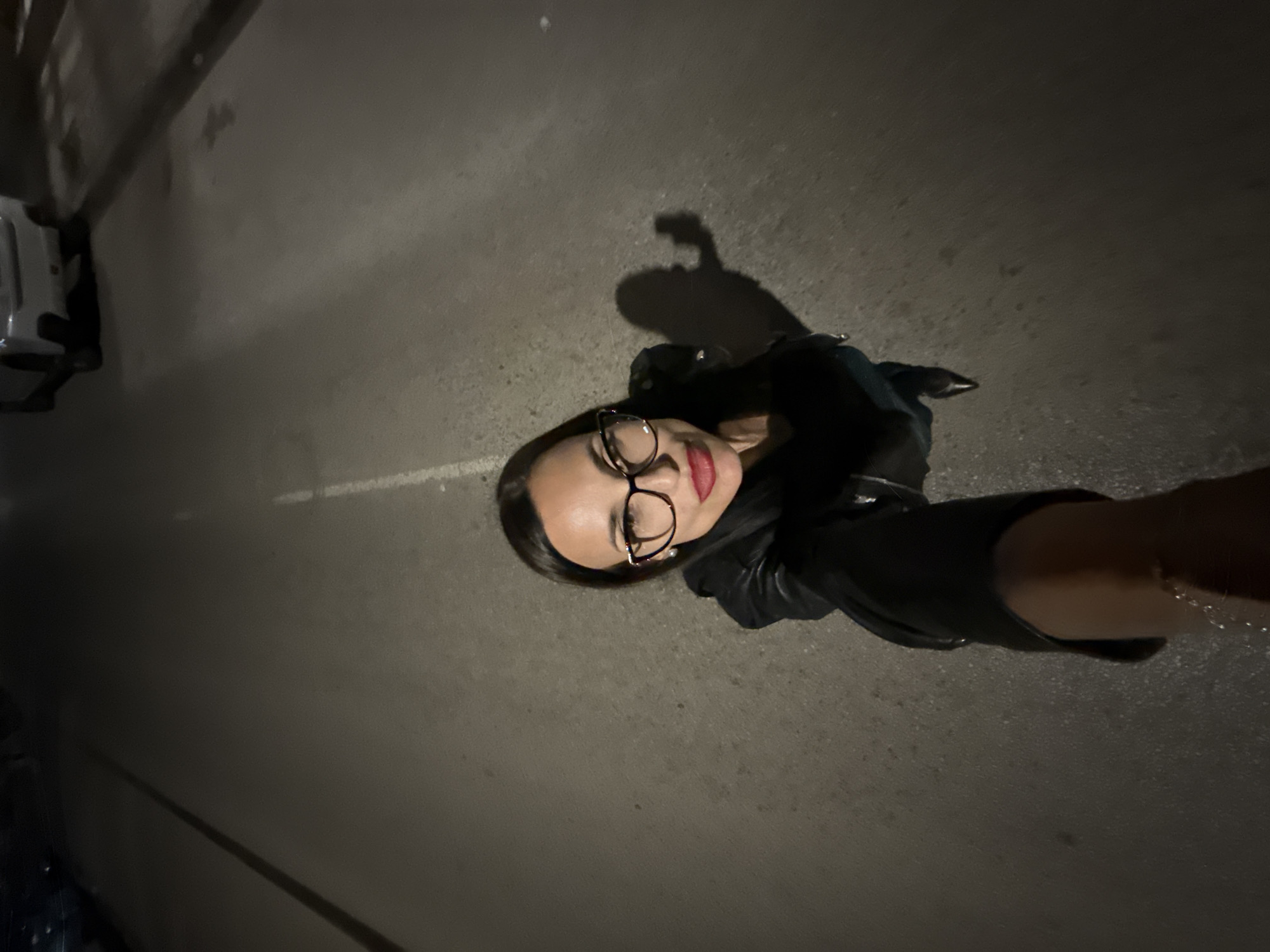 A woman is taking a selfie outdoors at night, standing on a gray asphalt surface with a white line marking on the left side. A blurred car is parked on the left, and the woman's shadow is visible behind her. She is wearing glasses, a black jacket, and has red lipstick. Her arm is extended to hold the camera. The location is Pleven, Bulgaria, but specific details are not possible from the image.