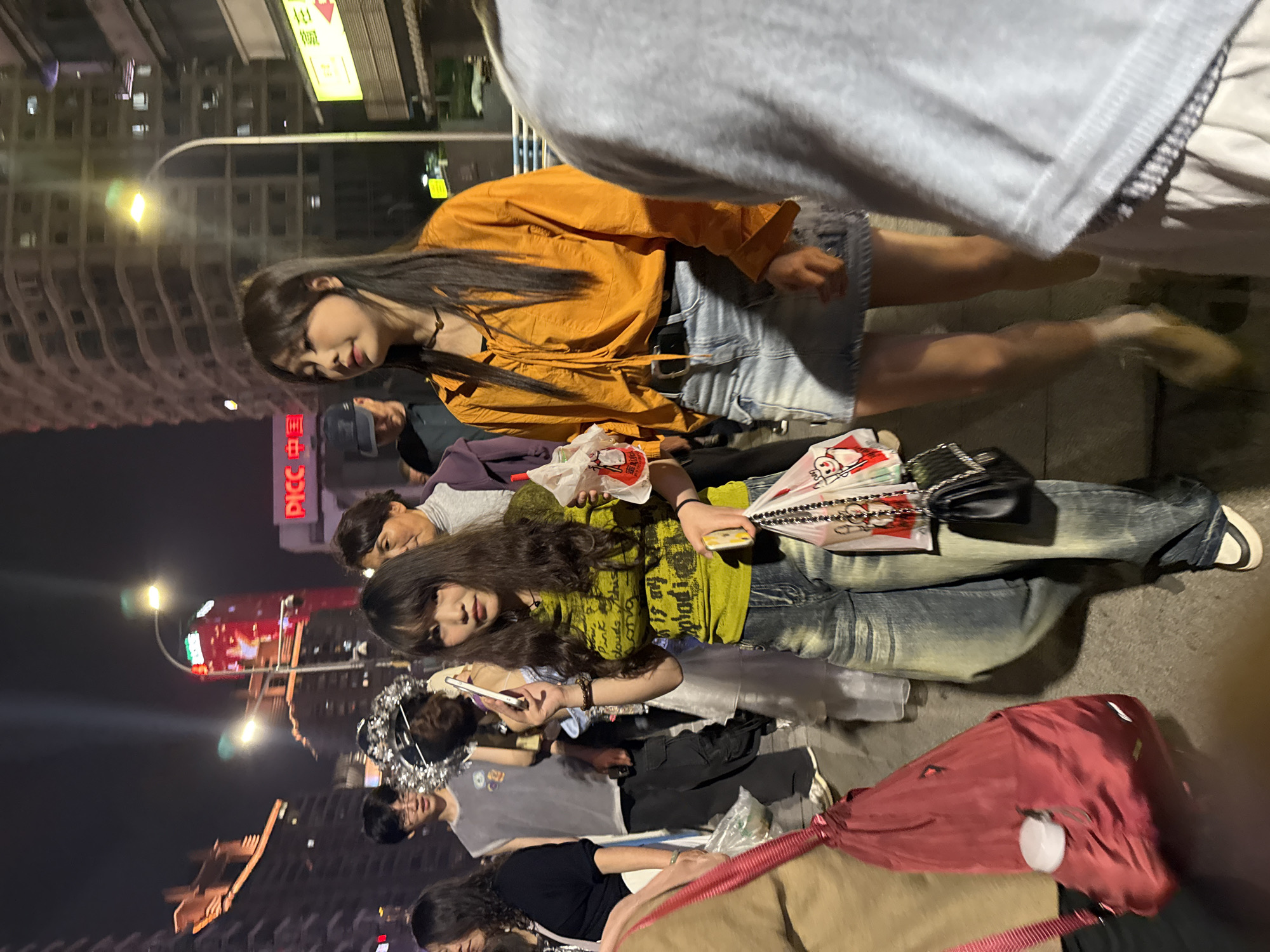 The image captures an outdoor scene at night in the city of Yuzhong District, China. Several individuals are present, possibly in a public gathering or a casual meetup.
One person is wearing an orange top and denim shorts. Another person has on a green shirt. There are also people dressed in casual street style.
The setting includes urban elements like buildings, streetlights, and what appears to be a bus stop or sign. Other visible objects are shopping bags, a red backpack, and mobile phones being used by some people. The overall atmosphere seems lively and informal, capturing an everyday scene in the city.