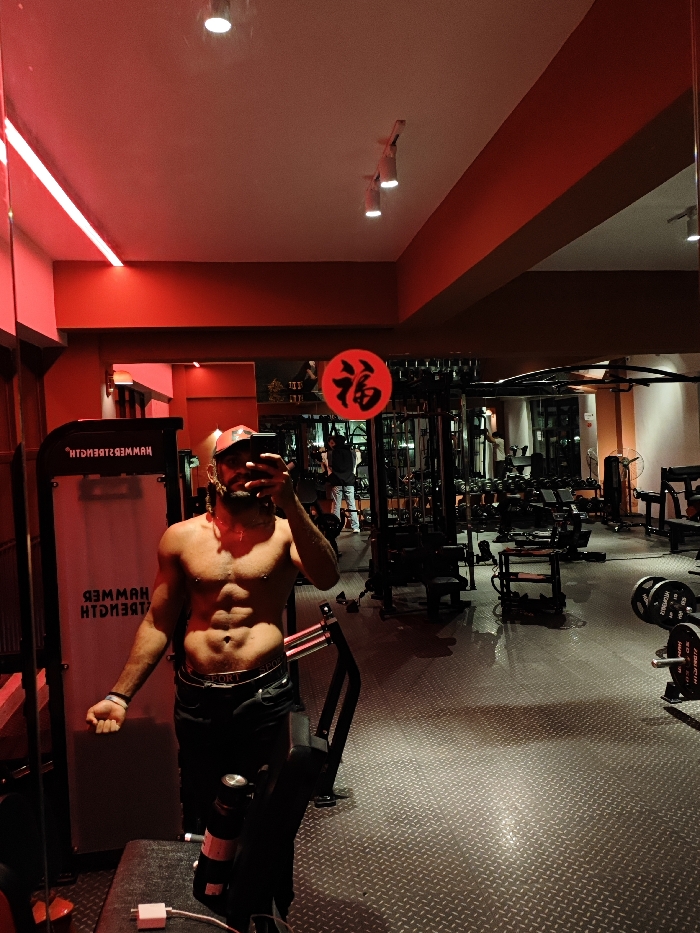 A man is captured in a mirror selfie inside what appears to be a gym or fitness center. He is shirtless, displaying a well-defined abdominal area, and is wearing black athletic shorts or pants. A cap is on his head, and he is holding a smartphone to take the picture, obscuring part of his face. His left arm is slightly flexed, showcasing his bicep.
The gym environment is characterized by dramatic red lighting, originating from LED strips running along the upper edge of a wall segment on the left and spotlights mounted on the white ceiling. The walls are predominantly painted in a deep red color, contrasting with the lighter ceiling.
In the background, various pieces of gym equipment are visible, arranged symmetrically due to the mirror's reflection. To the immediate left of the man, a black weight machine is prominent, with the mirrored text "HTGNERTZ REMMAH" (Hammer Strength) printed vertically in white. Further into the reflection, numerous other machines, weight racks, dumbbells, and benches can be seen. A red circular sign with a black Chinese character "福" (fú, meaning "good fortune" or "blessing") is prominently displayed above some equipment in the middle of the background. On the far right in the background, a large standing fan is visible.
The floor is covered with a dark, textured material featuring a diamond plate pattern. Reflections of the ceiling lights and possibly other light sources are visible on the glossy surface of the floor. Near the bottom of the mirror's reflection, a white cable (possibly a charging cable) and a black water bottle can be seen resting on a surface in front of the mirror. Another individual is partially visible in the deep background on the left side of the reflection, standing near some equipment. The overall atmosphere suggests an evening or night workout session, given the artificial and intense lighting.
