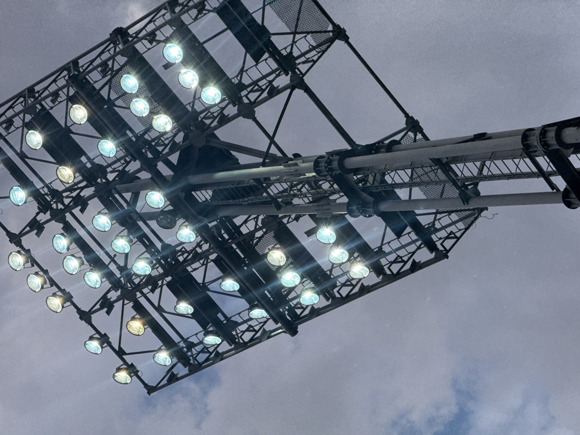 An outdoor shot looking up at a large metal stadium floodlight tower against a cloudy sky. The tower is angled, with numerous circular lights mounted on it, many of which are illuminated with a bright white or bluish-white light, creating a glare. The sky is overcast with patches of lighter clouds. The image does not provide any specific details or context related to the city of Abovyan, Armenia, or any information about people or specific activities occurring at this location. It is a static depiction of lighting equipment against a natural backdrop.