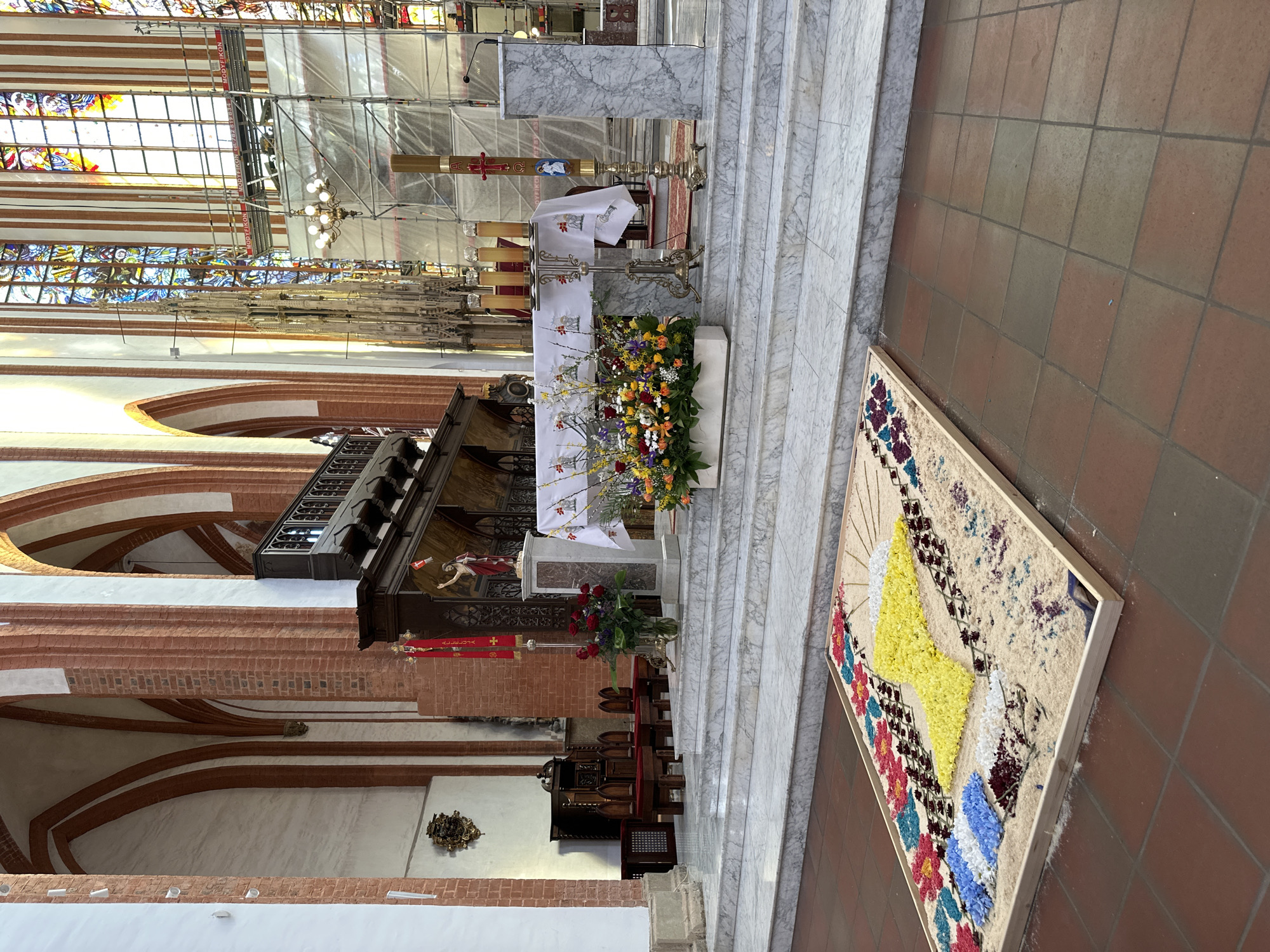 The image shows the interior of a church, likely in Wrocław, Poland. An altar is adorned with flowers, candles, and religious cloths. Scaffolding is visible near the stained-glass windows, indicating ongoing restoration or maintenance work. A large artwork made of what appears to be colorful grains or flower petals rests on the tiled floor near the altar steps. The architecture includes brick arches and marble steps.