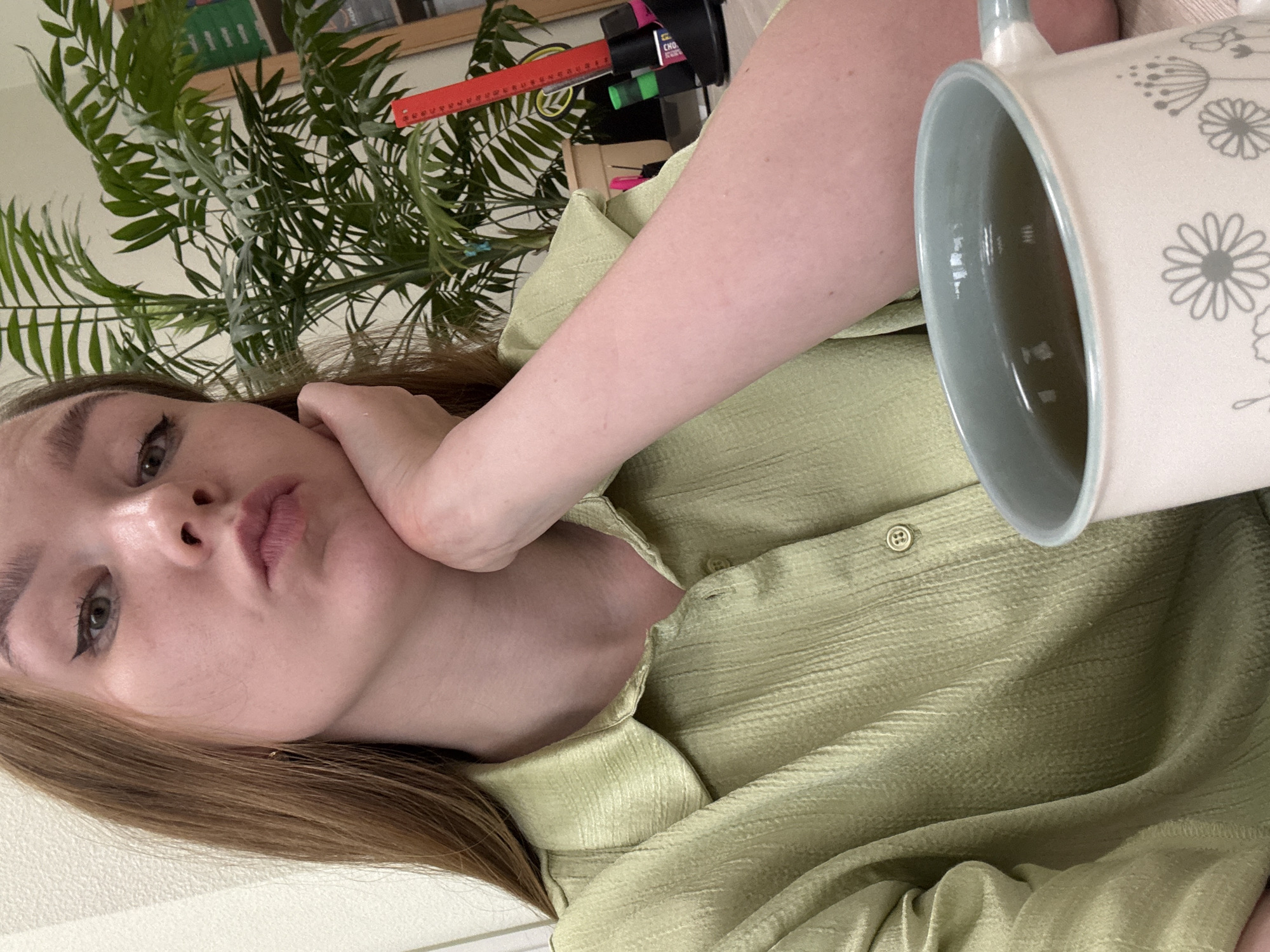 A person with light brown hair and green eyes is holding a mug with a floral design. The person is wearing a light green button-up shirt. A plant is visible in the background along with desk objects. The location is not discernible as Izhevsk, Russia.