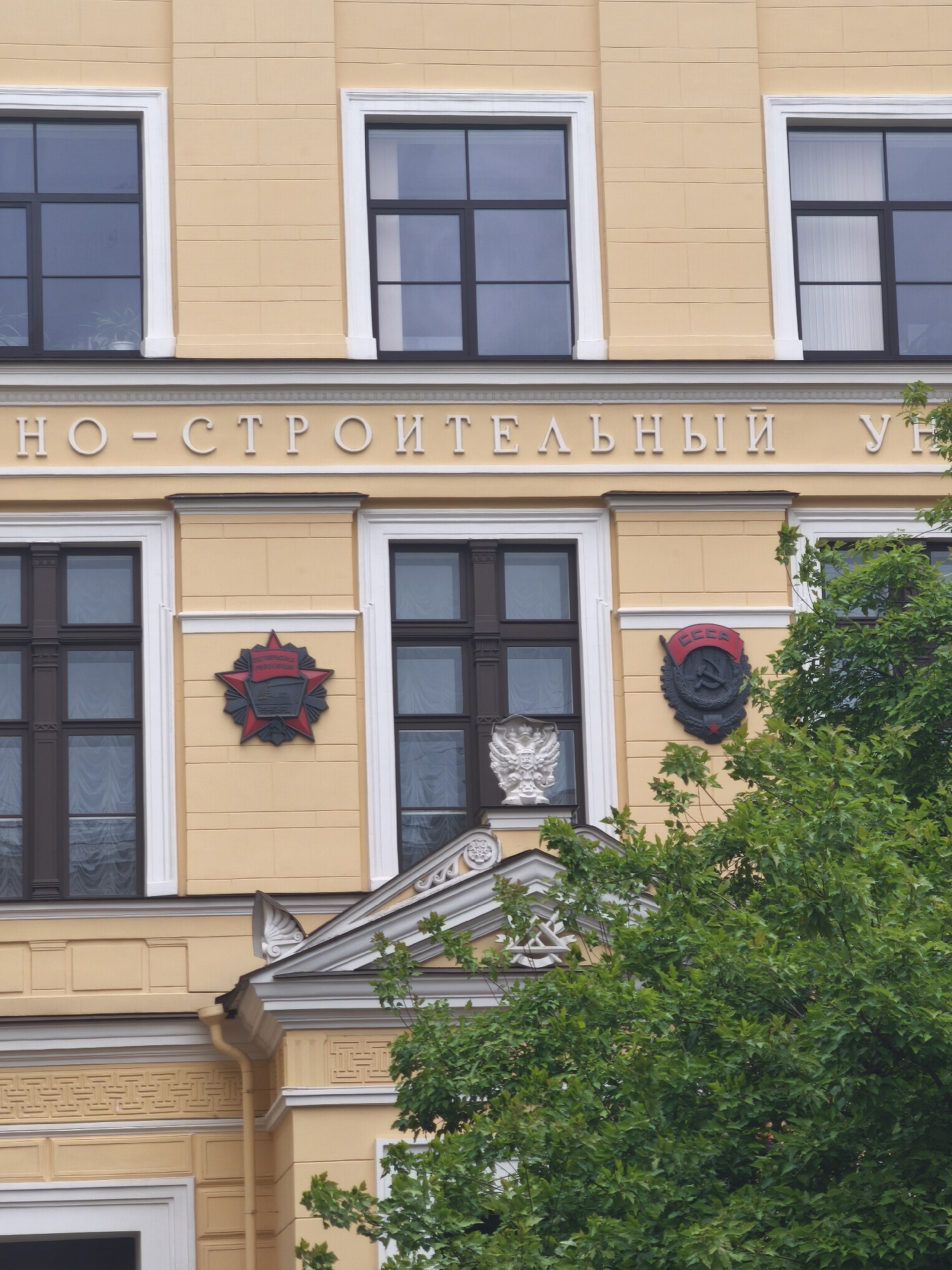 The image shows a section of a yellow building with architectural details, likely located in Saint Petersburg, Russia. The building has multiple windows with dark frames and white trim. Between the first and second-floor windows is an inscription in white Cyrillic letters, which is “МНО-СТРОИТЕЛЬНЫЙ УЧ.” Below the first-floor windows are Soviet-era emblems: a red star on the left and the hammer and sickle emblem of the Soviet Union on the right. Between them, is a double-headed eagle. These are mounted on the building's facade. Green foliage partially obscures the lower part of the building.