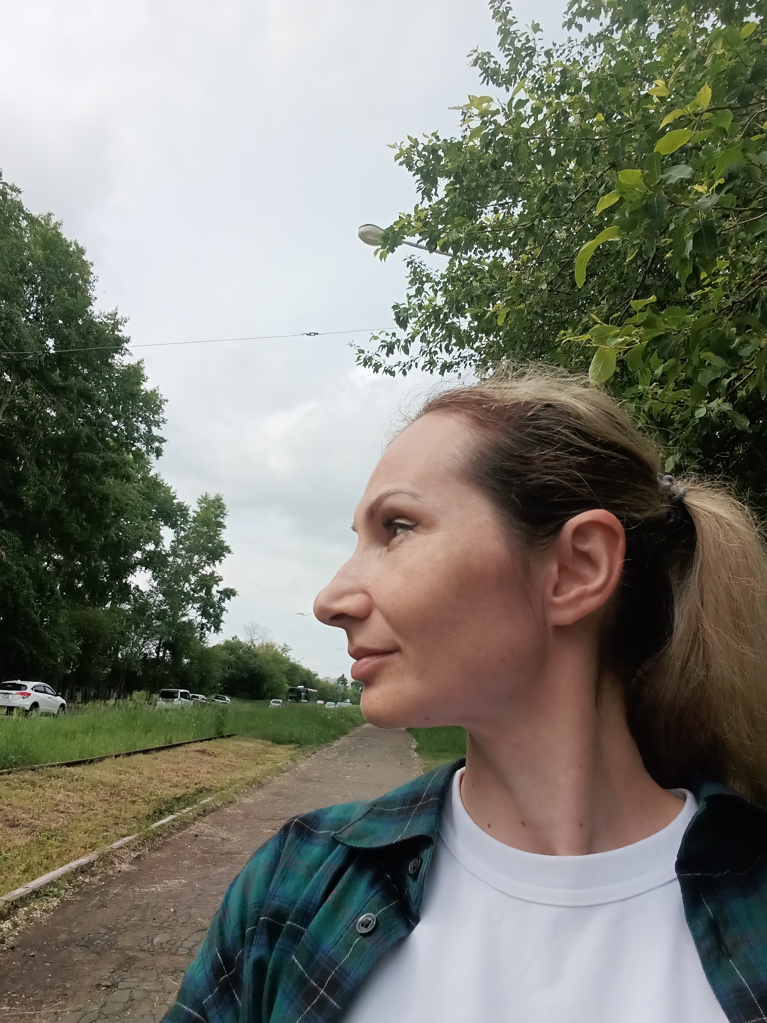 A middle-aged Caucasian woman with blonde hair pulled back in a ponytail, wearing a white t-shirt and a green and blue plaid shirt, is pictured in a close-up profile shot. She is looking to her left. The background shows a cloudy sky, trees, a dirt path, and a grassy embankment with a railway track. In the distance, several cars are parked along a road. The scene appears to be an outdoor area in Komsomolsk-on-Amur, Russia.