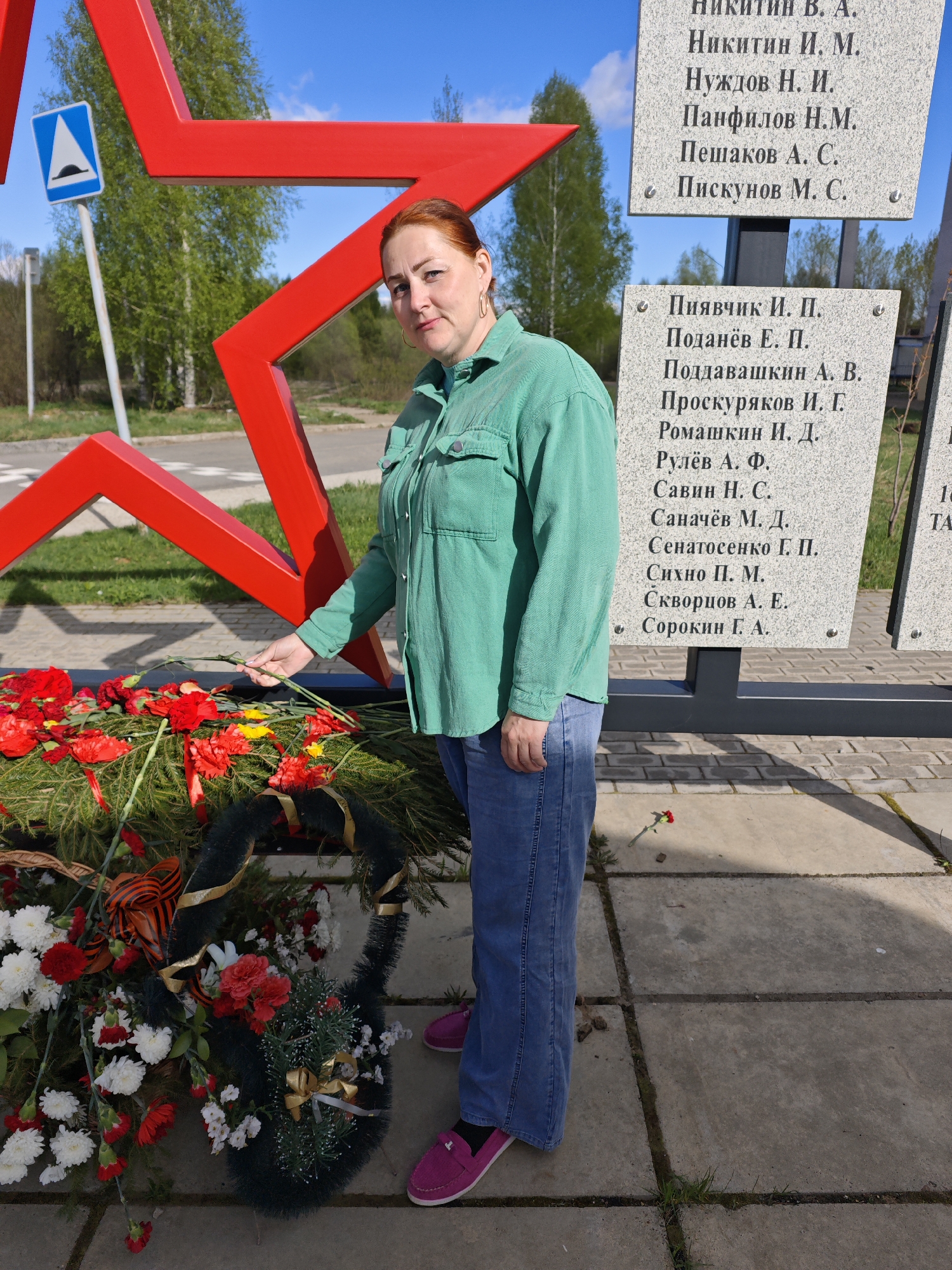 The image shows a woman standing in front of a memorial in Markovskiy, Russia. The memorial features a large red star and several stone plaques listing names. The woman is placing a red flower on a wreath adorned with red, white, and yellow flowers and a ribbon bearing black and orange stripes. She is wearing a green button-down shirt, blue jeans, and pink shoes. A road sign is visible in the background, along with trees and other greenery. The ground is made of concrete tiles with grass growing in the cracks.