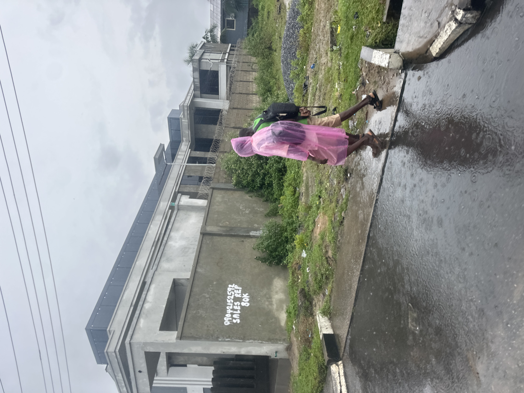 Two individuals are walking on a wet, paved surface in what appears to be the city of Badagry, Nigeria. The person in front is wearing a bright pink hooded raincoat and carrying a backpack. The second person, walking slightly behind, is wearing a green shirt and tan shorts, also carrying a backpack. They are walking away from the viewer, towards the right side of the image.
The environment is overcast, suggesting rain or cloudy weather, with visible raindrops creating ripples in the standing water on the pavement. Behind the individuals is a concrete wall with graffiti that reads "09042152572 SALES REP 80K." Beyond the wall, a modern, multi-story building with large windows and a flat roof is visible. The building's construction appears to be incomplete or in a minimalist style. Lush green vegetation, including bushes and some trees, grows around the building and along the roadside. A pile of dark gravel is also present in the background. The overall scene depicts an urban or semi-urban setting experiencing inclement weather.