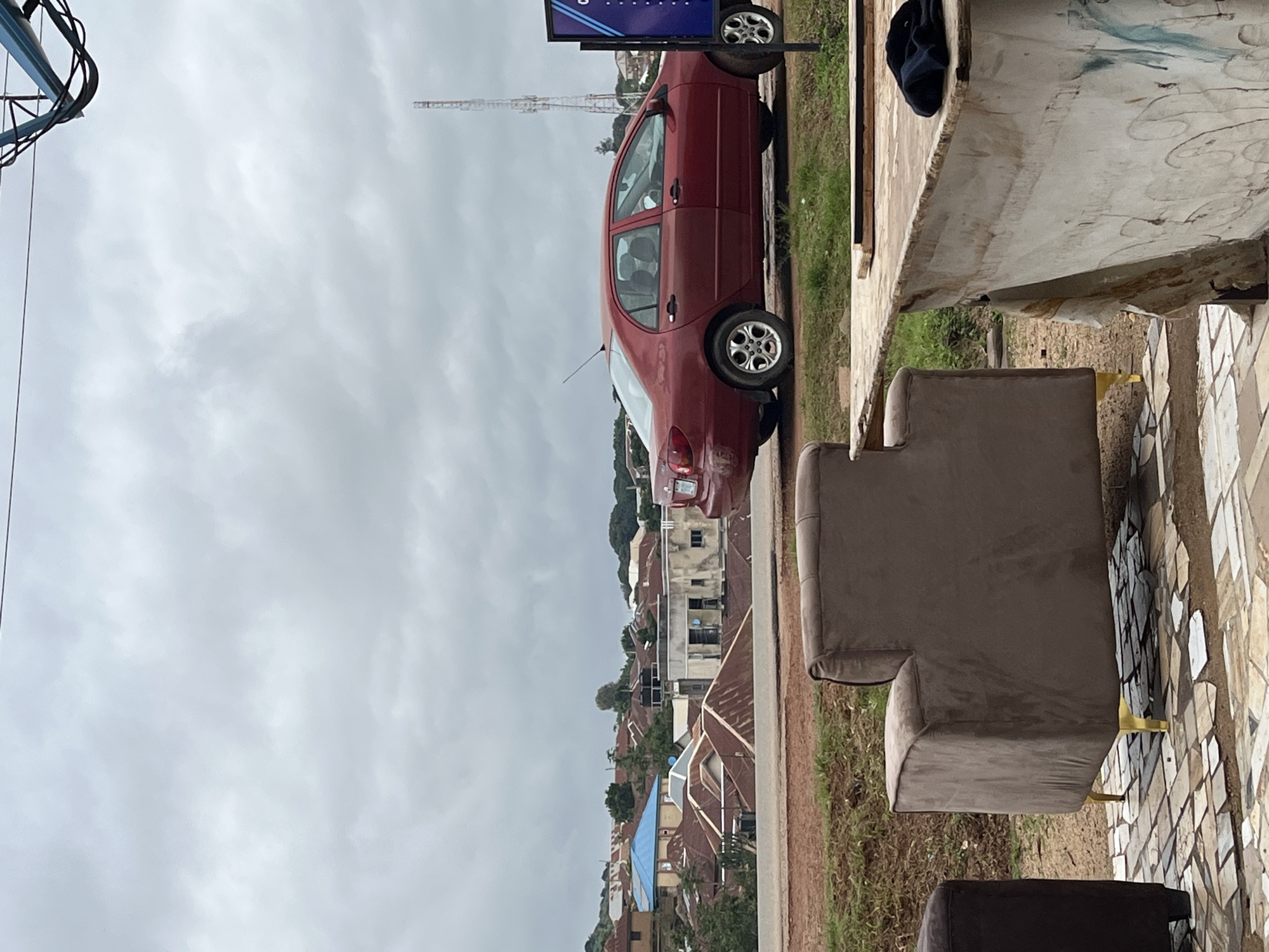 A red sedan is parked on a dirt road in an urban setting. The vehicle is facing right, with its rear end slightly angled towards the viewer. Behind the car, a dense residential area is visible, characterized by numerous buildings with corrugated metal roofs. In the distance, a tall telecommunications tower stands against a cloudy, overcast sky. In the foreground, two pieces of furniture are present: a brown upholstered armchair with gold legs on the left, and a section of a wooden counter on the right. The counter is draped with a stained and torn cloth. The ground is covered in patchy grass and patterned tiles. The scene appears to be located in Jos, Nigeria.