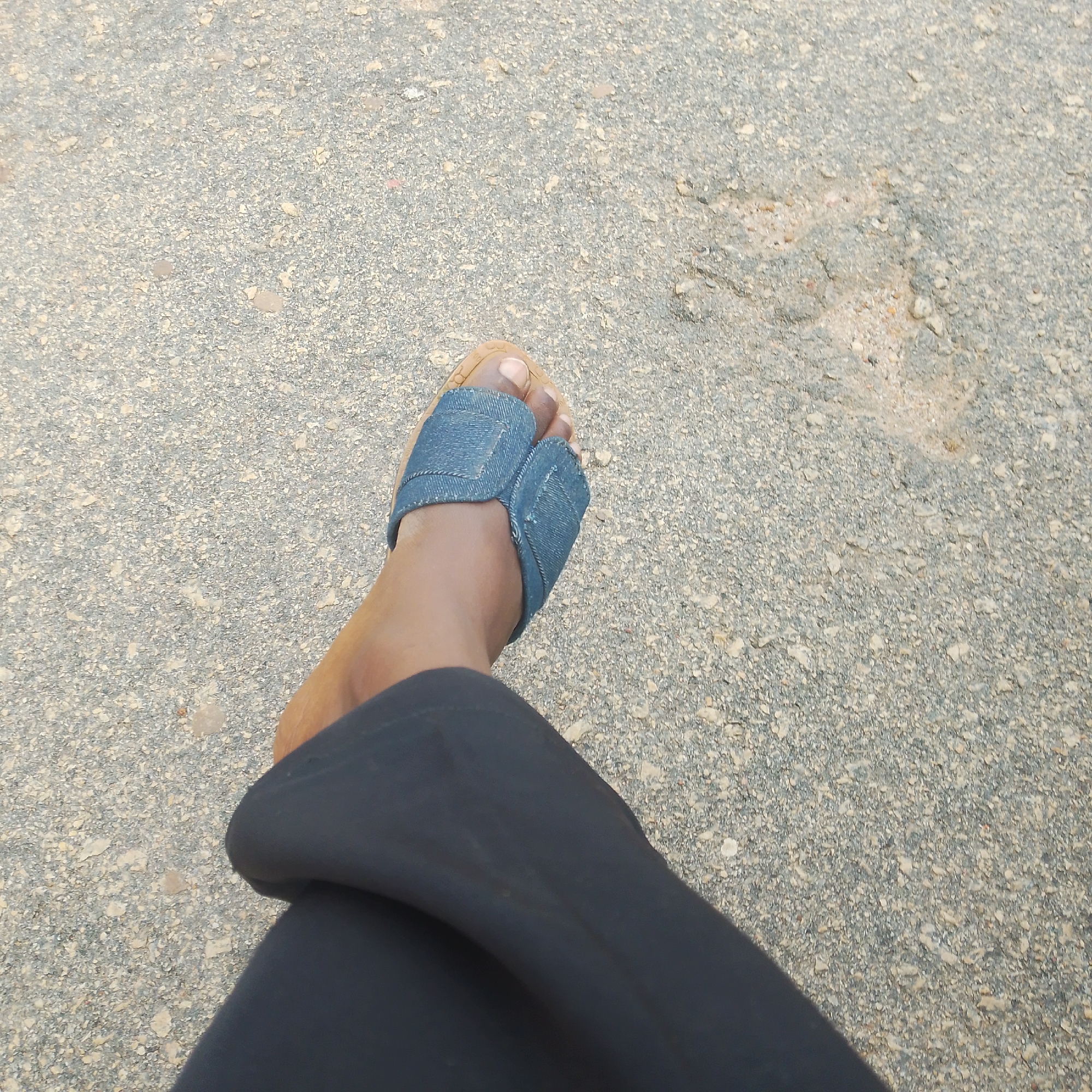 The image shows a close-up of a person's left foot wearing a denim flip-flop. The foot is resting on a grey, granular surface, likely asphalt or pavement. The person is wearing dark-colored pants that partially obscure the leg. The lighting suggests it is daytime, possibly outdoors. The flip-flop has a split design, with denim material covering the toes and the strap passing between the big toe and the second toe. The background is out of focus, showing the texture of the ground. There is no visible text in the image. The scene appears to be a casual setting in Bar Gada, Nigeria.