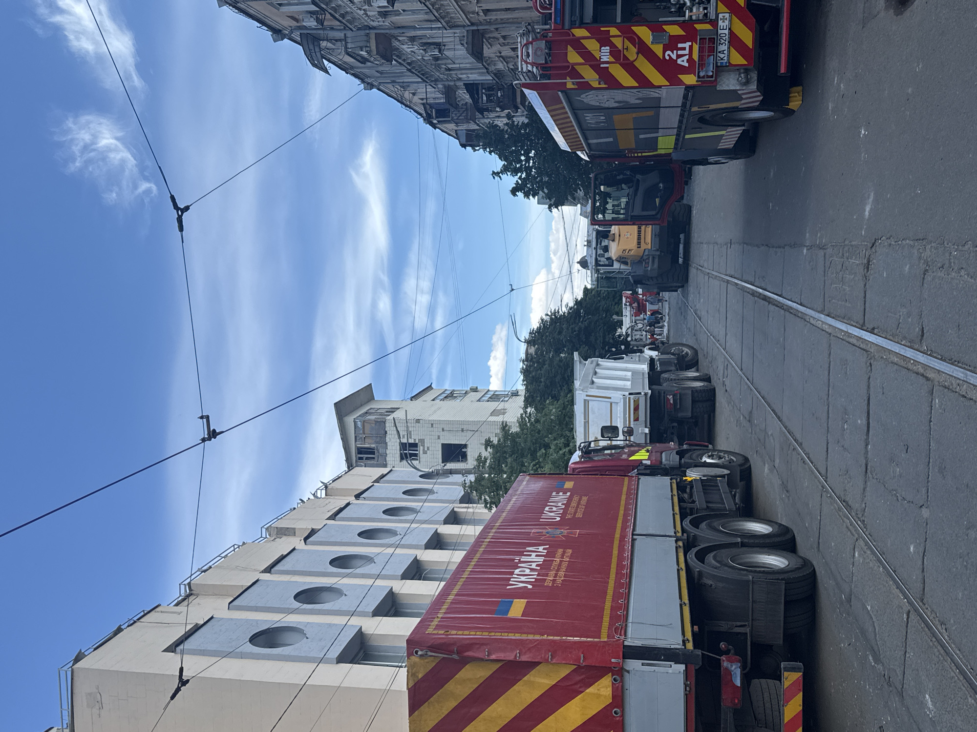 The image shows a street scene in Kyiv, Ukraine. Multiple vehicles are parked along tram tracks on a city street. At the front of the image is a large red truck with a tarp that has “Ukraine” printed in large letters, along with the Ukrainian flag. Behind the red truck, there is a white truck, a yellow construction vehicle, and a red fire engine. The vehicles are parked on a street that is lined on both sides with buildings. A taller building with a tower is visible in the background. Overhead tram cables run above the road. The sky is blue with scattered clouds.