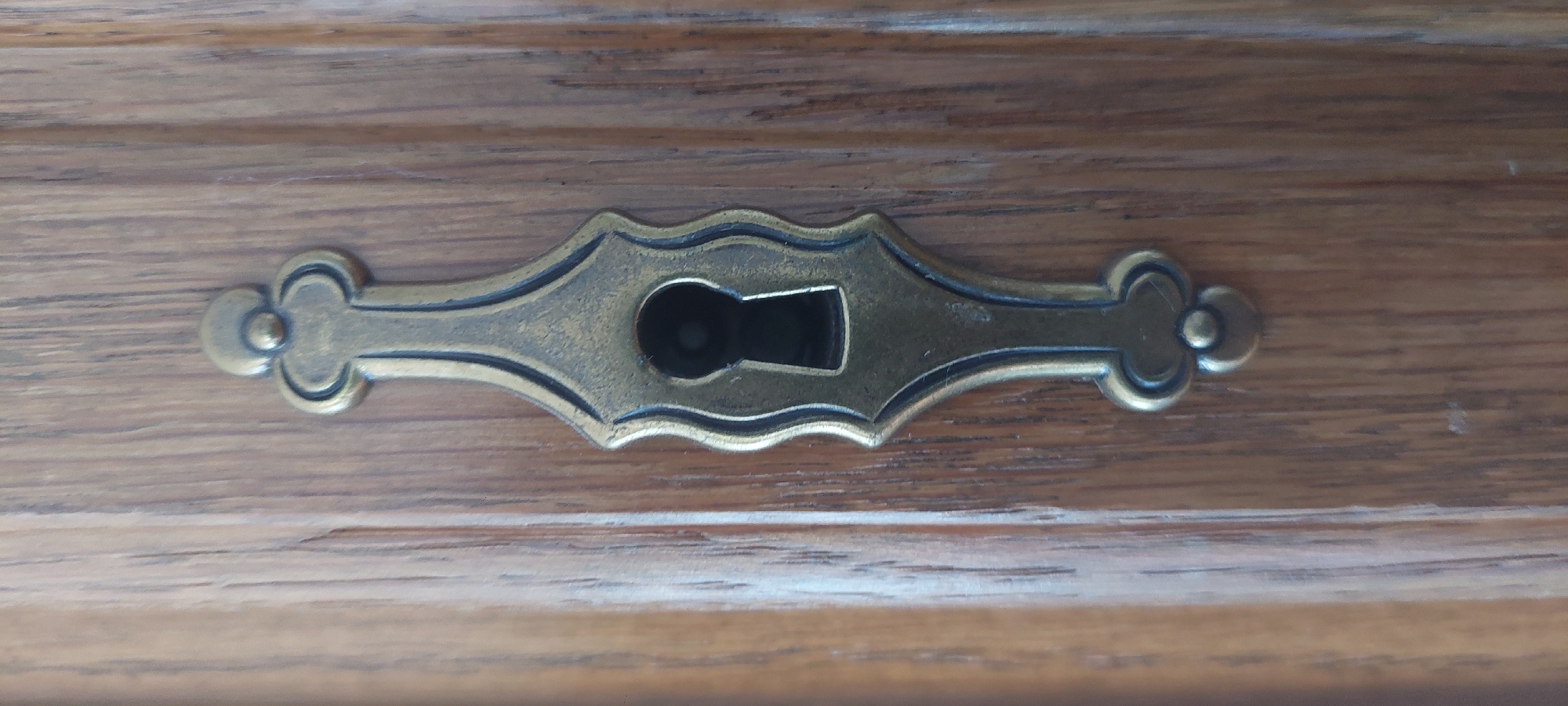 A close-up, vertical shot shows a brass keyhole plate on a wooden surface. The keyhole plate is ornate, with a decorative, elongated shape that tapers at the top and bottom and features curved edges. It is mounted on a wooden surface with a prominent grain pattern, suggesting it is part of a piece of furniture like a cabinet or door. The keyhole itself is dark, indicating it leads into a shadowed interior space. The lighting suggests an indoor setting with diffused light. The image is devoid of people or any activity. The setting is the village of Berod bei Hachenburg, Germany.