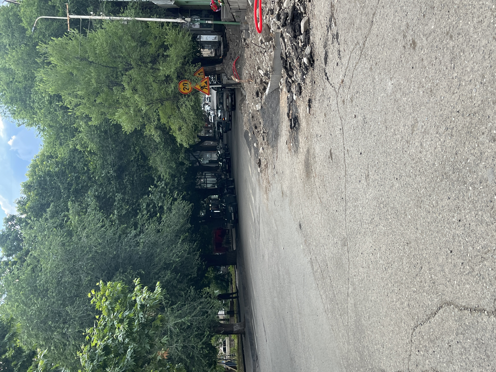 The image shows a street scene in Yeghegnadzor, Armenia, with construction or roadwork occurring on the right side of the road. A portion of the road surface appears to be torn up, with debris including asphalt, rocks, and concrete scattered along the curb. Red plastic piping is visible among the construction materials.
Several trees line the street, providing shade. Traffic signs are posted, including one indicating a speed limit of 20. Cars are parked and driving on the road. A person is standing near a tree on the left side of the street. Buildings are visible in the background. The sky is partly cloudy.