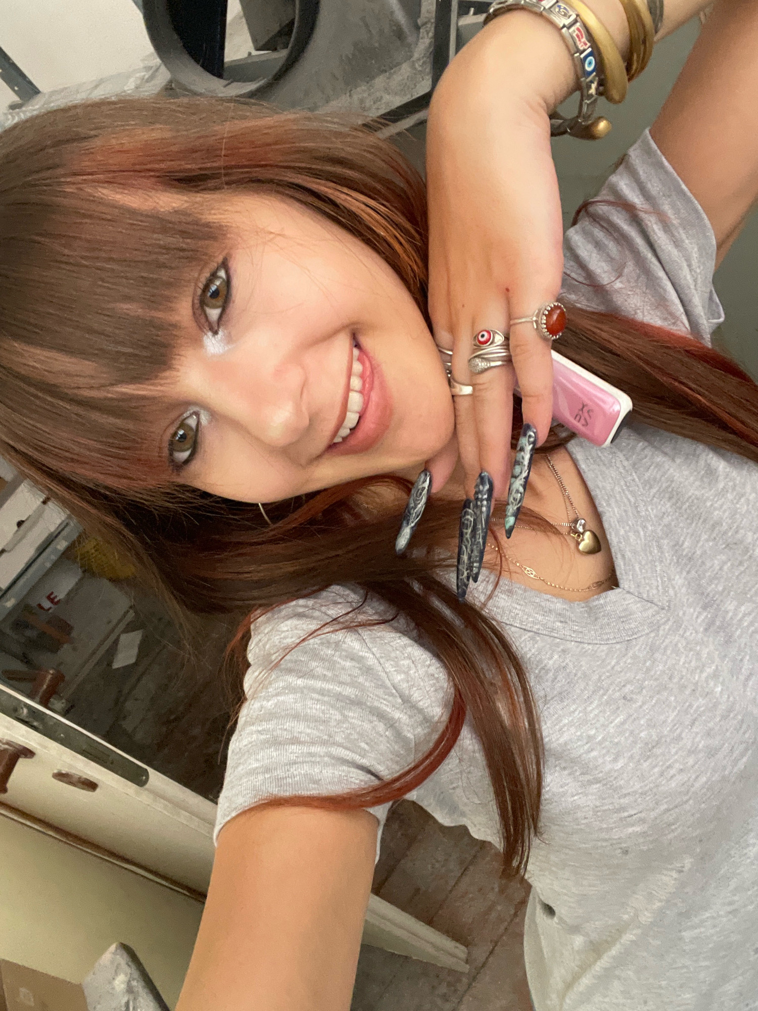 A woman with reddish-brown hair and bangs smiles at the camera in a selfie. She is wearing a grey t-shirt and several rings on her fingers, including one with a red stone and another with an evil eye design. Her nails are long and painted with a dark, marbled design. She is holding a pink object, possibly a vape, in her right hand. A gold heart-shaped pendant hangs from a thin chain around her neck. The background appears to be indoors, with a hint of industrial elements and some shelving or cabinets visible on the left. The lighting suggests it is daytime, possibly indoors with artificial light. There are no visible landmarks that would indicate the specific location within Raanana, Israel.