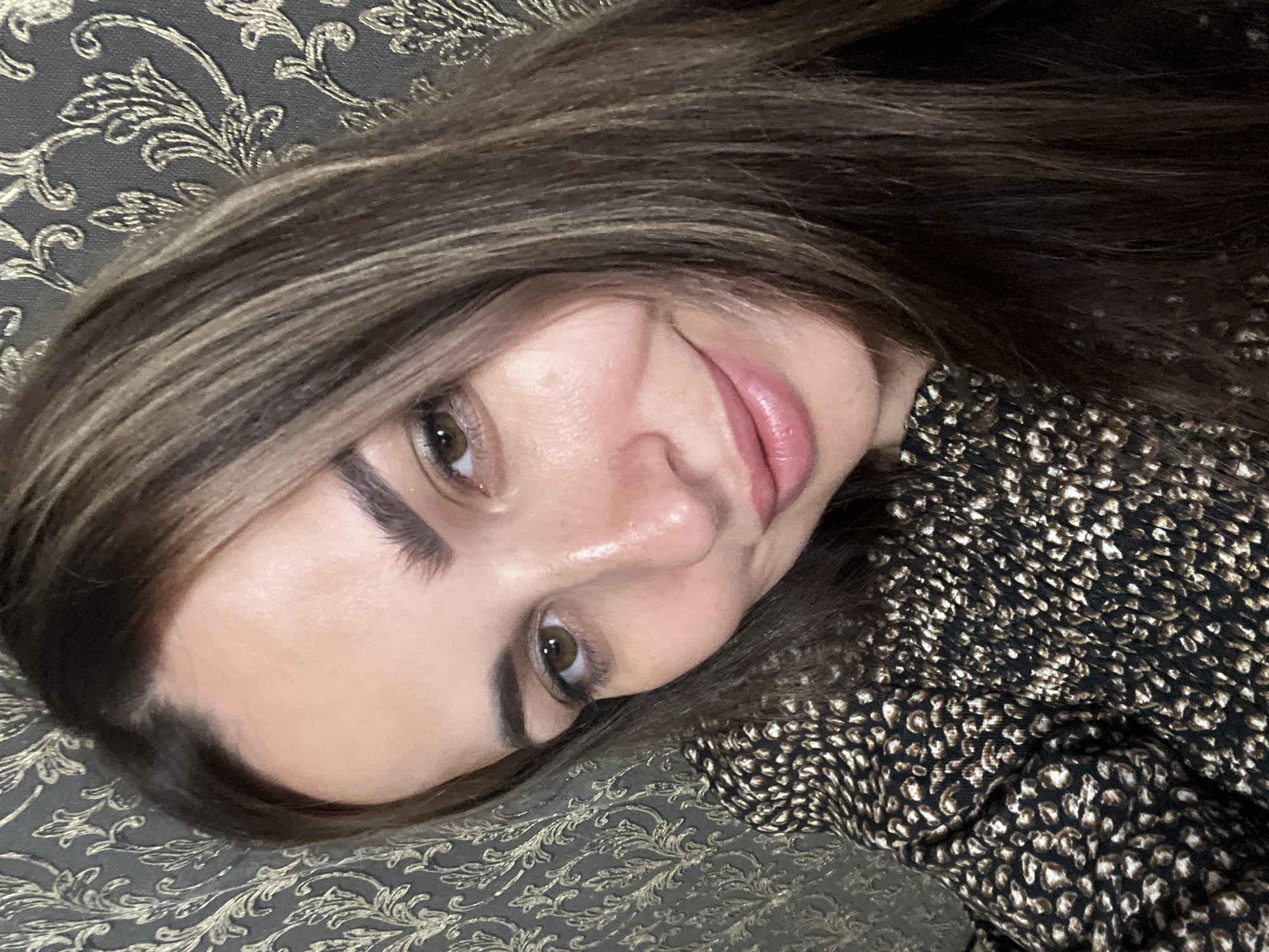 The media file contains a close-up, rotated portrait of a woman. She has long, dark brown hair with subtle highlights. Her right eye is visible, featuring a brown iris and dark eyelashes, and her eyebrows are well-defined. Her nose and lips are also visible, with her lips appearing to be slightly parted and colored with a pink lipstick. The woman is wearing a black top with a tan and white speckled pattern. She is lying down on a textured surface, possibly a couch or bed, with a dark grey fabric featuring an ornate, lighter grey or gold pattern. The lighting appears to be soft, suggesting an indoor setting, possibly during the day or evening. There are no visible signs of specific weather conditions or any text within the frame. The overall impression is a casual, relaxed pose, with the subject's face angled towards the camera.