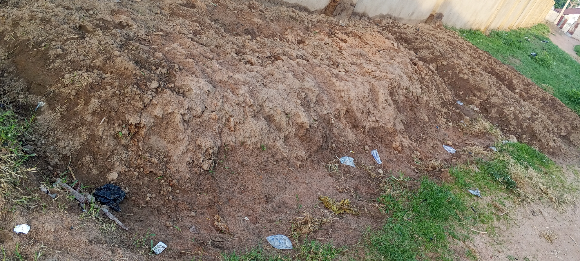 The image shows a large pile of excavated earth, possibly from construction or landscaping, in Jalingo, Nigeria. The pile is composed of brown soil and sand, with some dry vegetation and scattered pieces of trash, including plastic bags and wrappers. A section of a concrete wall is visible in the background, along with some dry grass and what appears to be a dirt path. The overall scene suggests an outdoor area that may be undergoing development or has been recently disturbed. There are no people visible in the image.