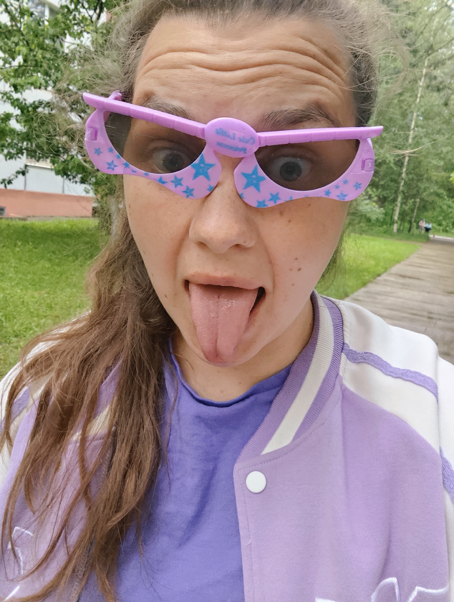 A young woman is captured in a close-up, playful selfie, sticking out her tongue and making a wide-eyed expression. She has light brown, wavy hair framing her face and across her shoulders. Her forehead shows slight wrinkles from the raised eyebrows, emphasizing the humorous mood.
She is wearing large, novelty sunglasses that are purple with light blue star patterns. The frames are unusually shaped, covering the upper part of her face more than typical sunglasses, and they feature small decorative elements above each lens. Visible text on the top frame of the sunglasses reads "Our Trip" with a heart symbol.
Her attire consists of a bright purple t-shirt layered under a white and light purple varsity-style jacket. The jacket has white stripes along the collar and shoulder seams, and a prominent white button is visible on the front.
The setting is outdoors, appearing to be a park or green area, possibly during daytime with soft, natural lighting. In the background to the left, green foliage from trees and parts of a multi-story building are visible. To the right, a wooden or paved pathway extends into the distance, lined by more trees and greenery. Two indistinct figures can be seen walking further down the path, suggesting a public recreational space. The overall atmosphere conveyed is lighthearted and candid.