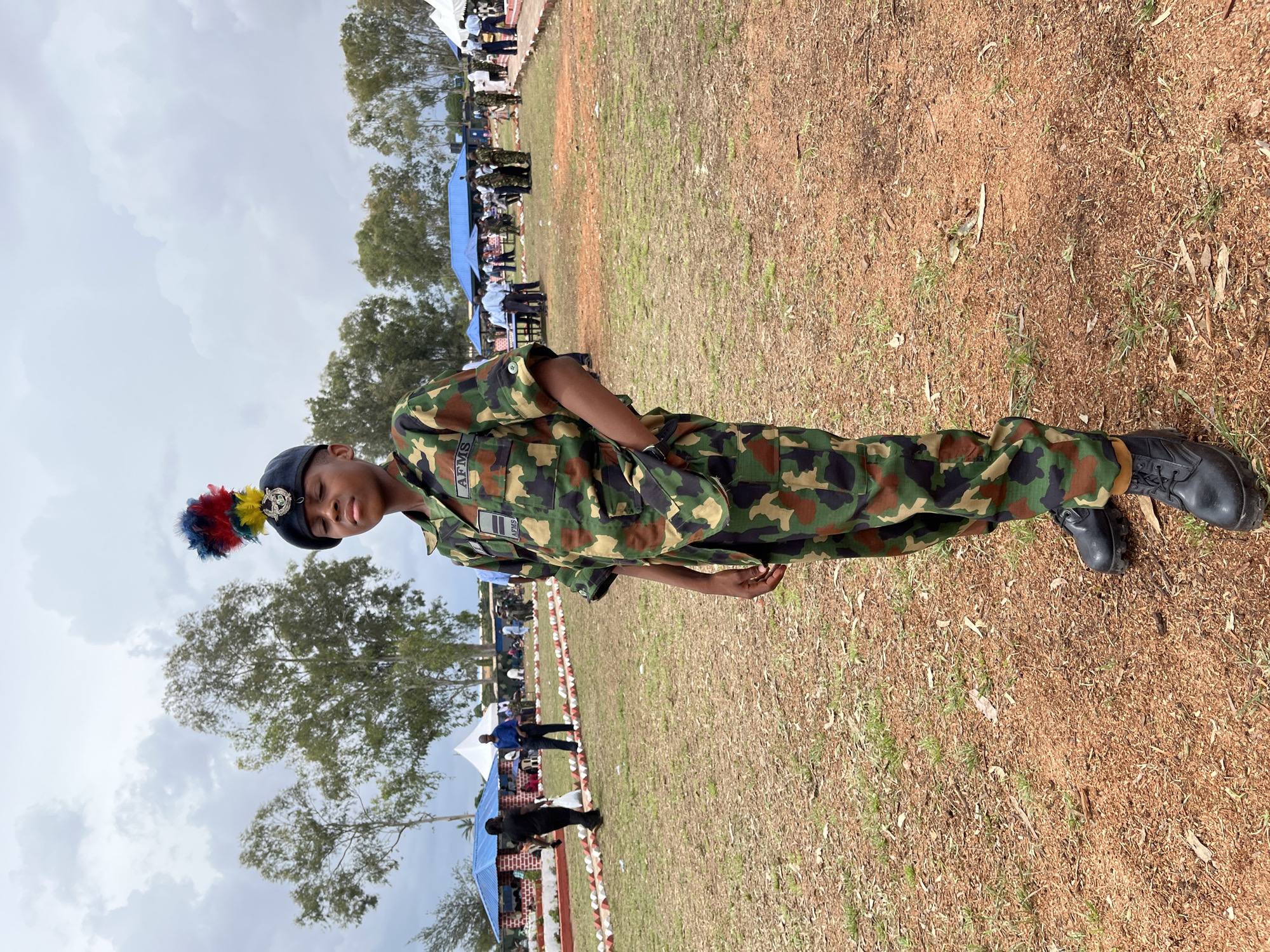 A young person in a military uniform stands in a field. The uniform is camouflage with green, brown, and black patterns. They are wearing a dark beret with a colorful plume. The person has their hands in their pockets and is looking down. In the background, there are tents, people in civilian and military attire, and trees. The ground is covered in dry grass and reddish-brown dirt. The sky is cloudy. The scene appears to be an outdoor event or gathering in Barakin Lerawi, Nigeria.