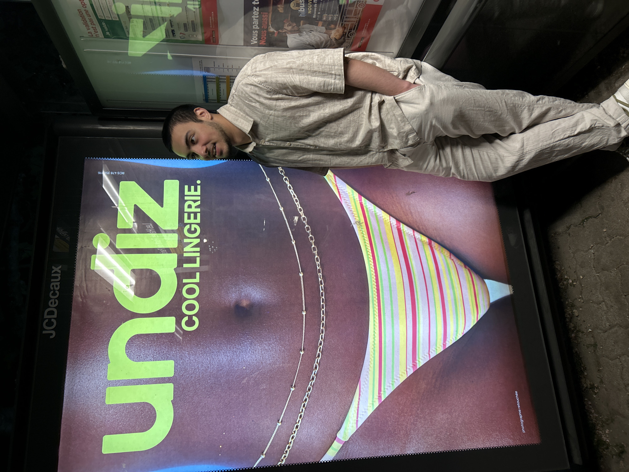 A light-skinned male with short, dark hair, light facial hair, wearing a light-colored shirt and pants, poses for a photo next to an advertisement for undiz, a lingerie company. The advertisement features a close-up of a female torso wearing a striped bikini bottom and body chains. The advertisement is placed in a JCDecaux display case. A second advertisement, including the phrase "Vous partez to" and transportation information for Strasbourg, France, appears above the lingerie advertisement. The ground appears to be concrete.