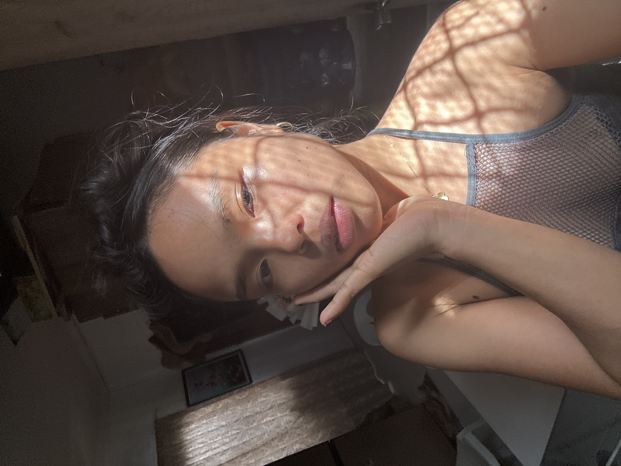 The image depicts a young woman taking a selfie in an indoor setting, likely a bedroom or living space. Sunlight streams through a window or other opening, casting distinct shadows across her face and body. She has her hair pulled up in a messy bun and is wearing a gray, mesh-like top. Her left hand is raised, touching her cheek. The background is dimly lit and appears cluttered, with a curtain visible on the left and what looks like shelves or stacked items on the right. The overall impression is of a casual, perhaps early morning or late afternoon shot, with natural lighting playing a significant role. There are no identifiable landmarks or cues within the image that would definitively place it in Davao City, Philippines.