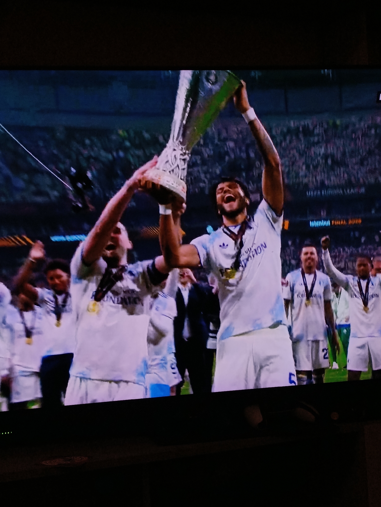 The image depicts a celebration, likely related to a sports victory. Several individuals are seen wearing white jerseys with medals around their necks, indicating they are members of a winning team. Two players in the foreground are hoisting a trophy above their heads, while others in the background are raising their arms in jubilation. The backdrop reveals a large crowd gathered in what appears to be a stadium. A sign reading "UEFA EUROPA LEAGUE Istanbul FINAL 2020" can be seen in the background.
The image cannot be placed in the city of Plovdiv, Bulgaria, as the sign indicates the final took place in Istanbul.