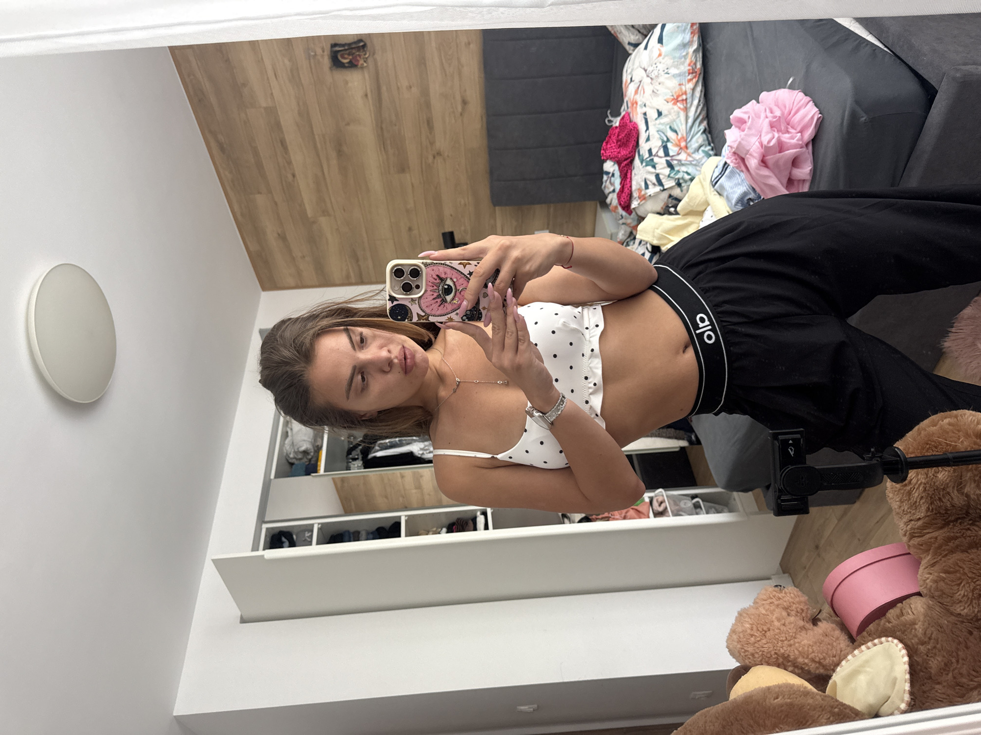The image is a self-portrait taken in a bedroom. A young woman with brown hair is standing in front of a mirror, holding a smartphone. She is wearing a white polka-dot bralette and black loose-fitting pants with "alo" branding on the waistband. The bedroom appears somewhat cluttered, with clothes on the bed and an open closet in the background. A large teddy bear and a pink hat box are visible to the left of the frame. The room has a wooden-paneled wall behind the bed and a white ceiling with a circular light fixture. There is no identifiable information in the image that would indicate the location is specifically in Plovdiv, Bulgaria.