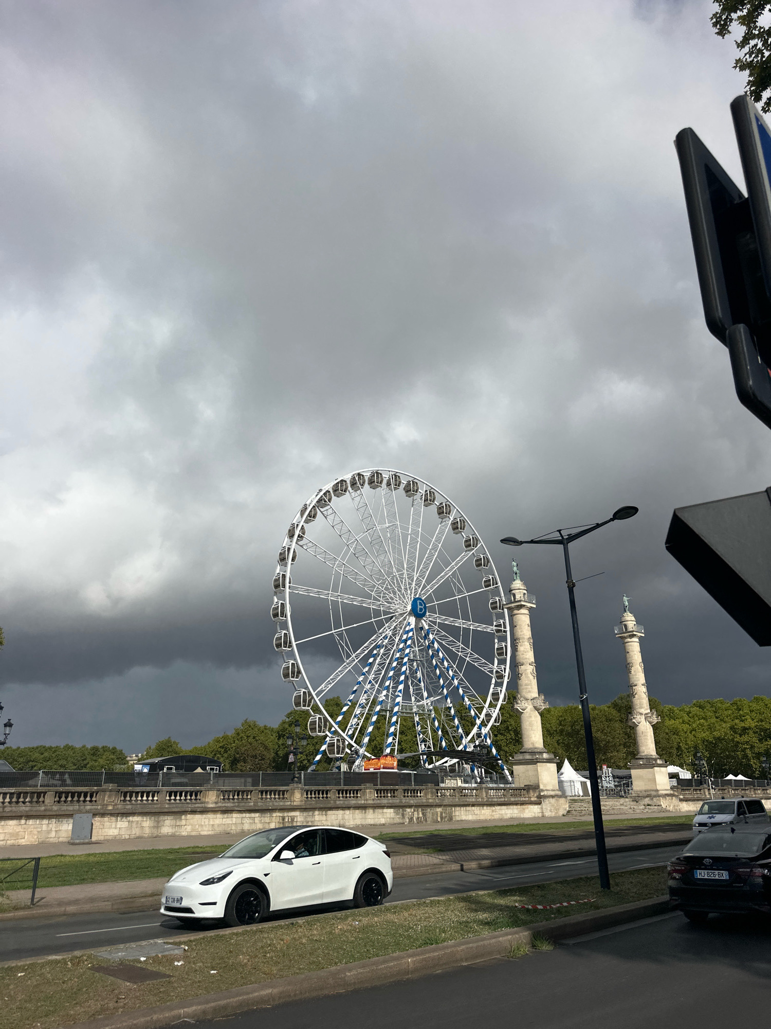 The image captures a scene in Bordeaux, France, featuring a large Ferris wheel with white spokes and blue accents against a dramatic, stormy grey sky. Two tall, ornate stone columns topped with statues stand to the right of the Ferris wheel. In the foreground, a white electric car, identified by its license plate as a Tesla Model Y, is parked or moving on a street. Behind the car and the Ferris wheel, a stone embankment with balustrades separates the road from a treeline. The sky is overcast with dark, ominous clouds dominating the upper portion of the image, suggesting impending rain.