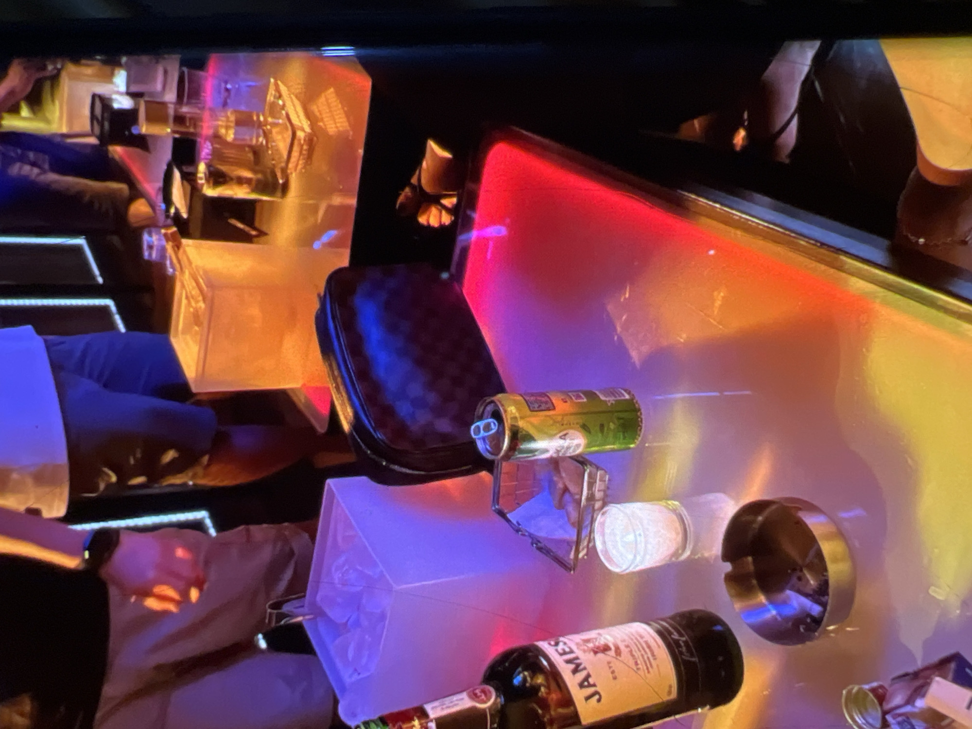 The image depicts a dimly lit bar scene in Lagos, Nigeria. In the foreground, a bottle of Jameson whiskey stands next to a small, clear shot glass. To the right of the shot glass, a can of beer is partially visible, and behind it, a small wire basket holds a few food items. An ashtray, containing cigarette butts, sits in the lower left corner of the frame. In the background, a white ice bucket filled with ice is prominent. The table surface is reflective, with colored lights from the bar creating vibrant reflections. Two individuals are partially visible; one on the left is wearing beige shorts and a dark t-shirt, with a smartwatch on their wrist. Another individual on the right is wearing dark trousers and a white t-shirt. The overall ambiance suggests a social gathering or a night out in a bar.