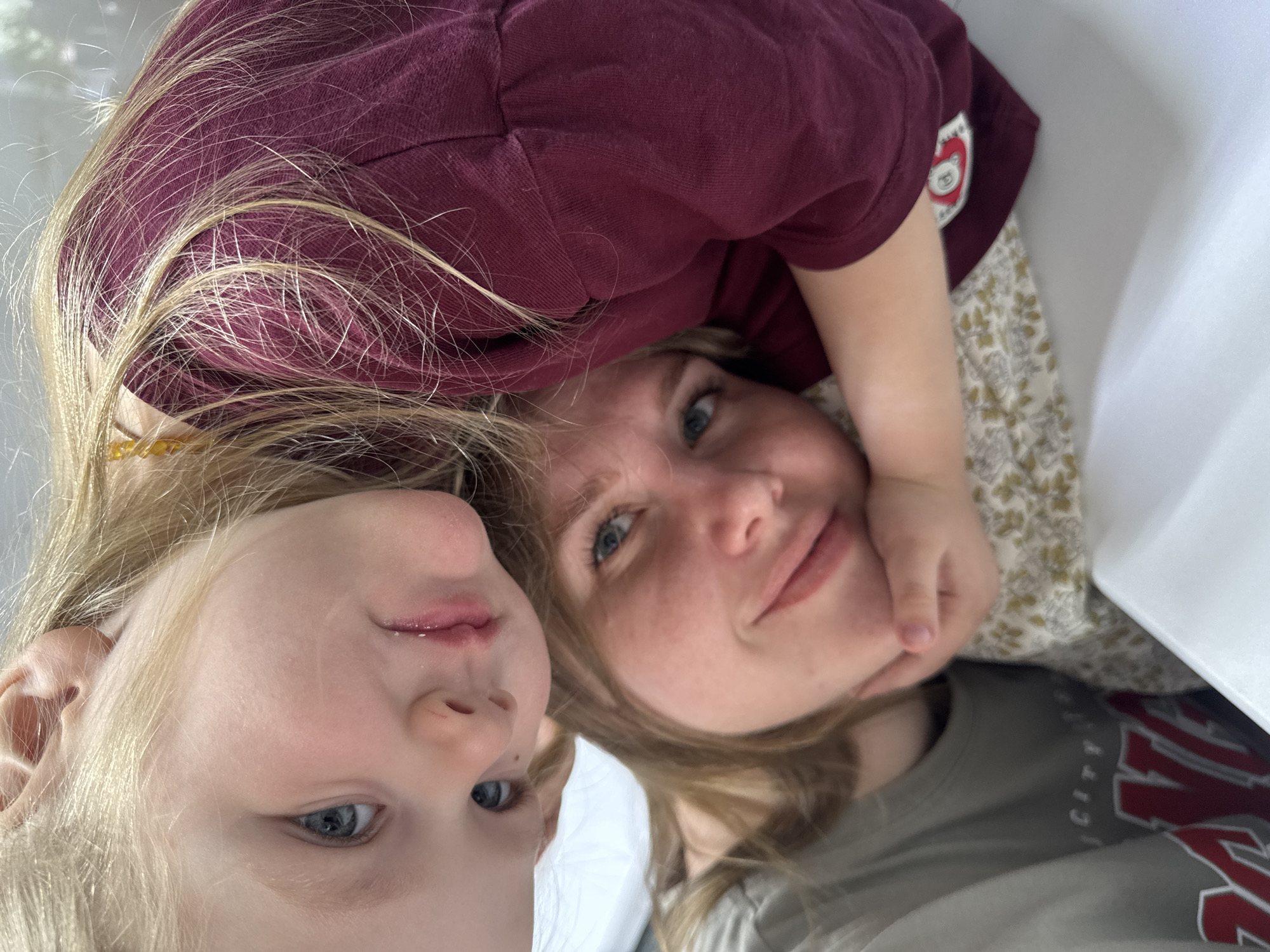 The image depicts a selfie of a woman and two children. The child on the left has blonde hair and blue eyes. The woman has blonde hair and blue eyes and is looking directly at the camera. The other child, wearing a burgundy shirt with a patch, is lying on the woman. The woman is wearing a gray t-shirt. The background is blurry and white. The location is likely indoors. The image was taken in Gornyi (Dvurechenskoe S/P), Russia.