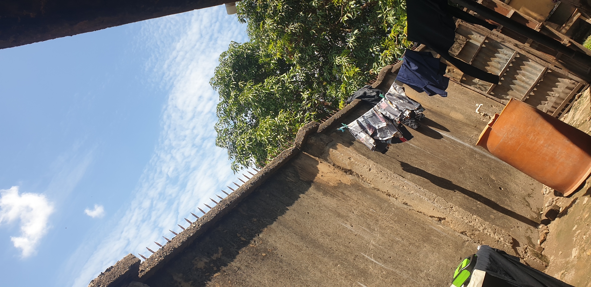 This image captures an outdoor scene in Sabon Tasha, Nigeria, viewed from a low, tilted angle. A weathered concrete wall, topped with a row of rusted anti-climb spikes, extends diagonally from the bottom-left to the upper-left of the frame. Direct sunlight casts prominent, elongated shadows on this wall and the uneven earthen ground below. Several articles of clothing, including patterned garments, a black jacket, and dark blue trousers, hang from a clothesline stretched across the concrete wall. A large, orange plastic barrel rests on the ground at the bottom-center, positioned beneath a small white spigot protruding from the wall. To the right, partially visible, is a structure constructed from corrugated metal sheets. Above and behind the concrete wall, a dense canopy of green trees is present. The upper portion of the image displays a clear blue sky with scattered white clouds, and two small, dark aerial specks are visible. No people are depicted in the scene.