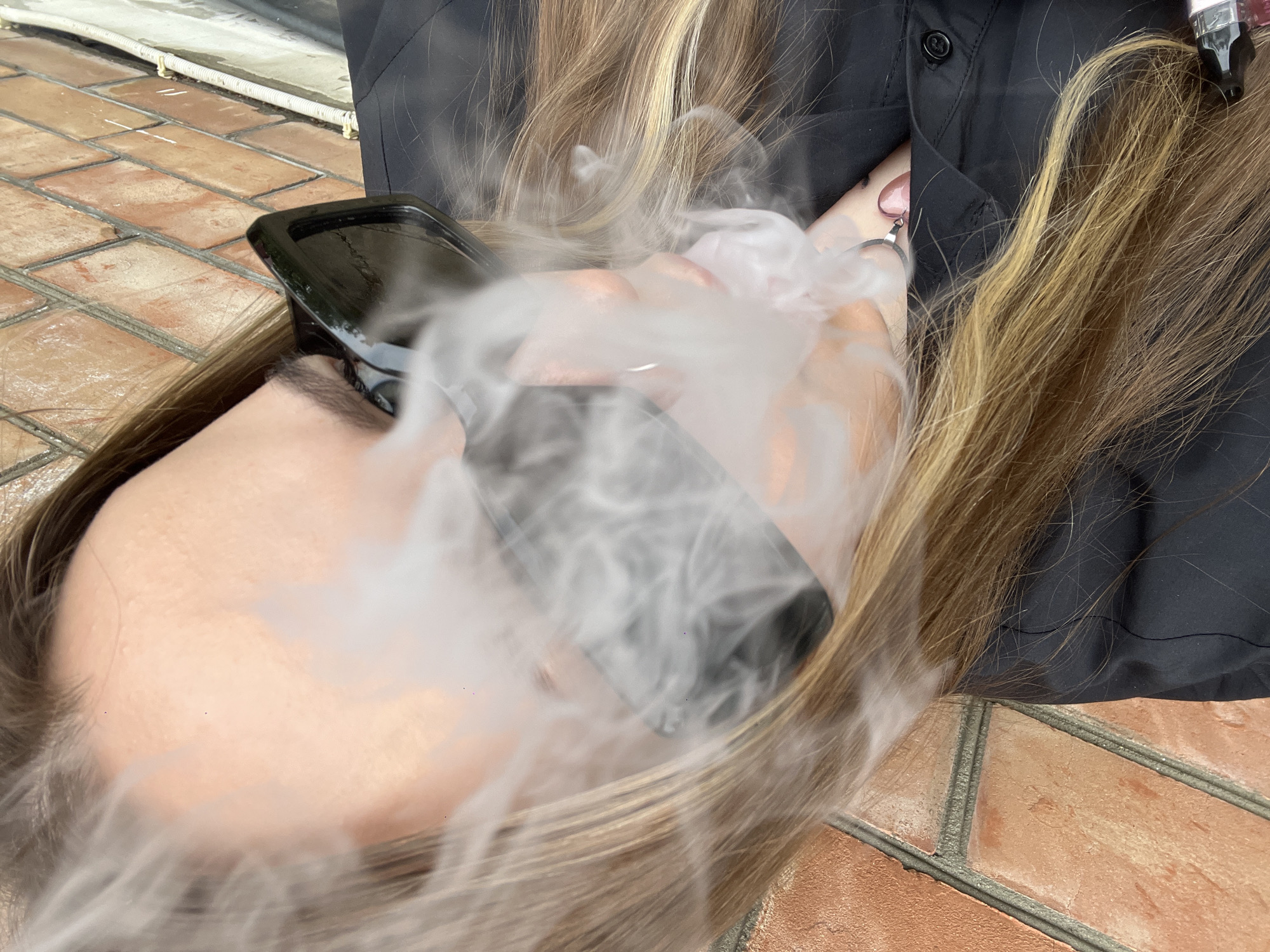 A person with long blonde hair is vaping, producing a large cloud of vapor that obscures their face and the phone they are holding. They are wearing a black jacket. The background shows a brick surface, suggesting an outdoor setting, possibly a patio or walkway. The location is Novopokrovskaya, Russia, as indicated by the prompt. The person is holding the phone close to their face, and vapor is billowing around it.