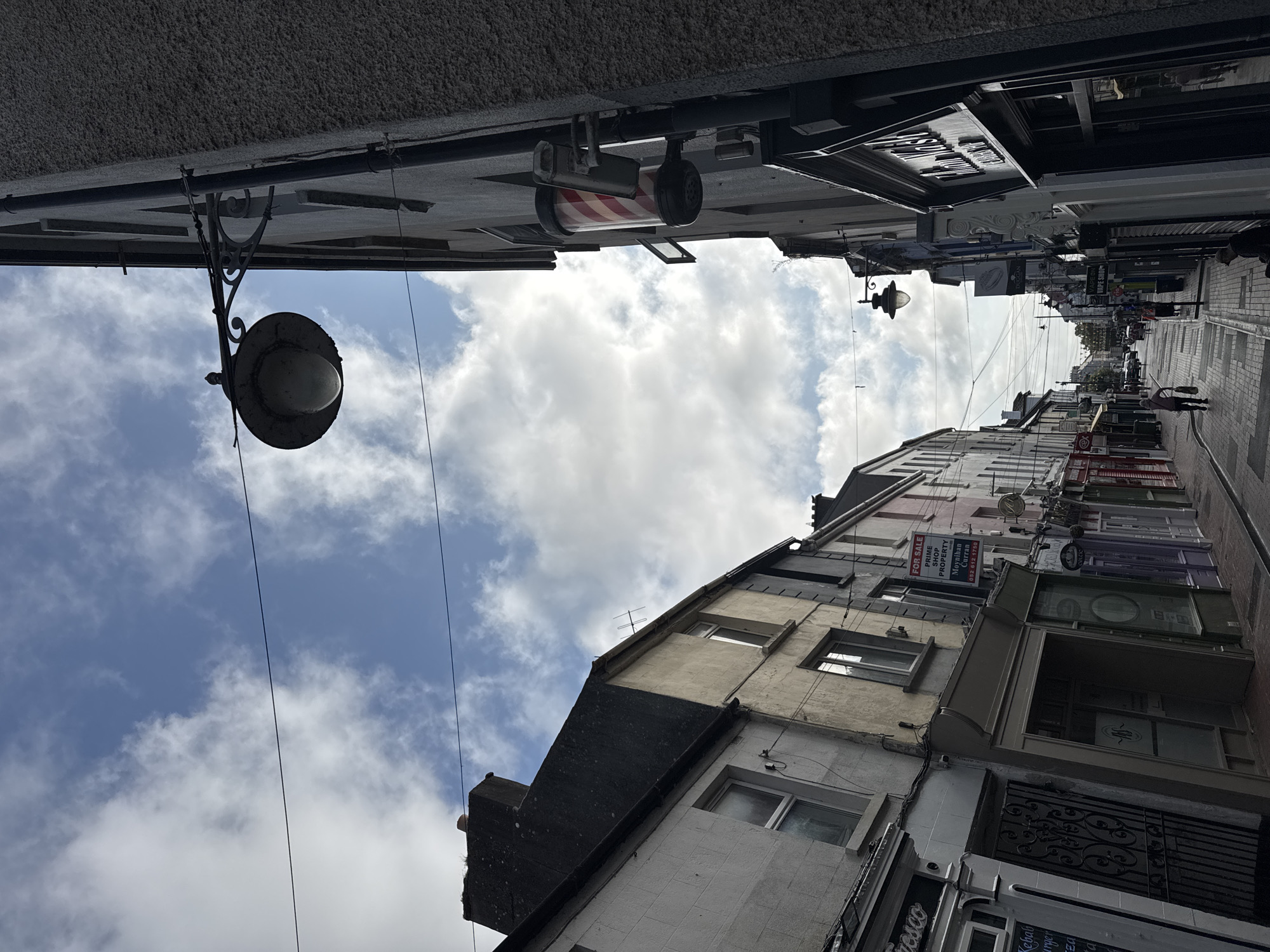 The image shows a street scene in Clonmel, Ireland, looking down a narrow street lined with shops. The sky is partly cloudy with patches of blue. Overhead, telephone or power lines are strung across the street.
On the right side of the street, a barber pole is mounted on the side of a building. Above the barber pole, a security camera is visible. Further down the right side of the street, a sign for "Thom Waite" is visible on a shopfront.
On the left side of the street, a row of shops is visible, including one with a sign that says "Fresco" and another with "Kebab burger." A "FOR SALE" sign is hanging from one of the buildings on the left.
A few people are walking down the street, and cars are parked further down the road. The street is paved with brick or cobblestone. The overall impression is of a typical urban street in a historic town.