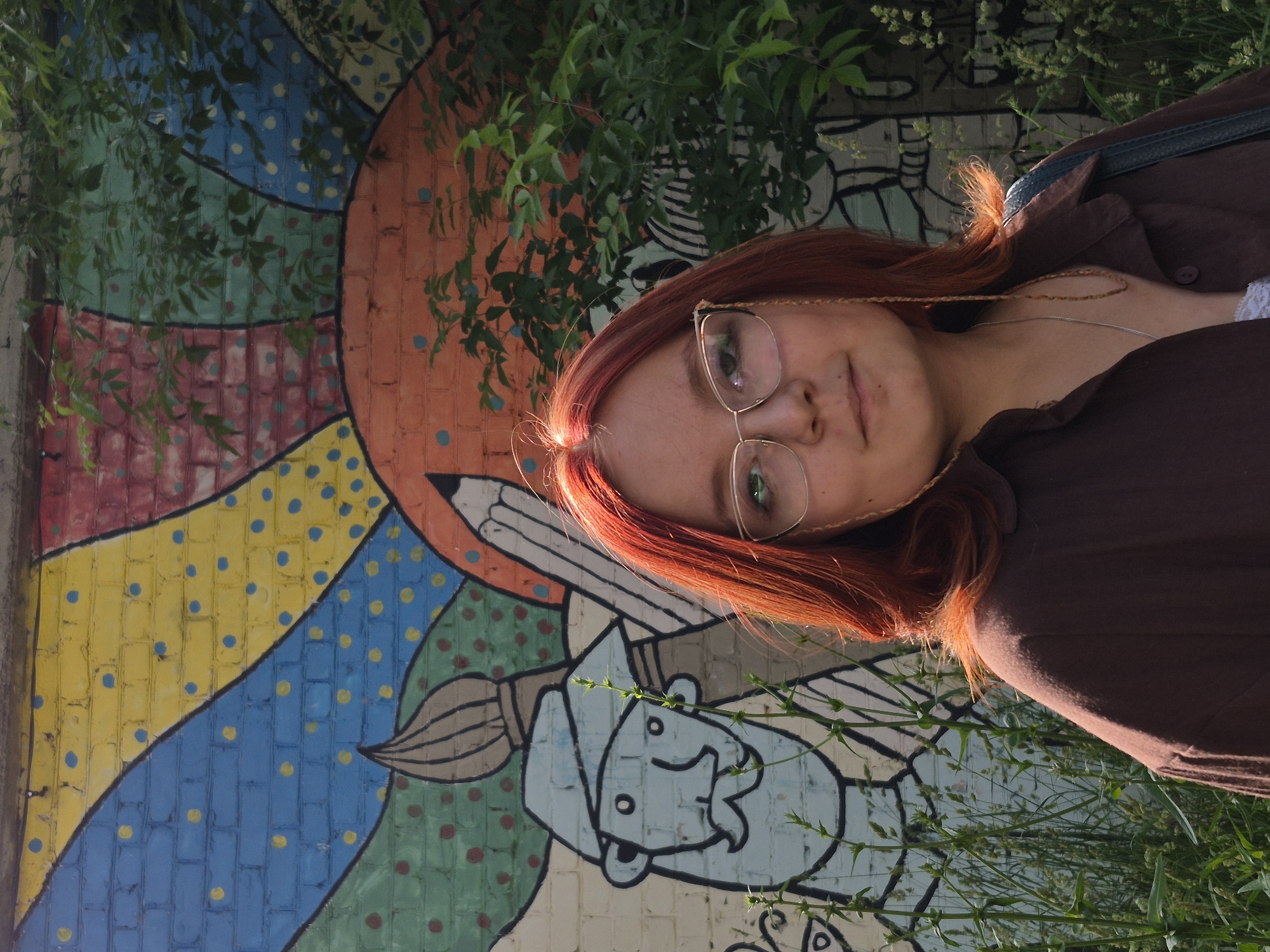 This is a vertical, eye-level, medium shot of a young person with vibrant red hair, wearing glasses and a brown shirt, leaning against a colorful mural on a brick wall. The mural features abstract shapes and patterns in shades of orange, blue, yellow, pink, and green, with polka dots. A large, stylized paintbrush and a pencil are also depicted in the artwork. Green foliage hangs from above and to the left of the person, partially obscuring the mural and creating a dappled light effect. The person's face is turned towards the camera, and they have a slight smile. Their eyes are visible through their glasses. A thin, braided necklace is visible around their neck. The background of the mural appears to have some graffiti or a continuation of the artwork with black lines. The presence of green plants suggests an outdoor setting, possibly a park or a street with greenery. The lighting appears to be natural, indicating it is daytime. The overall mood is casual and relaxed. There is no visible text in the image.