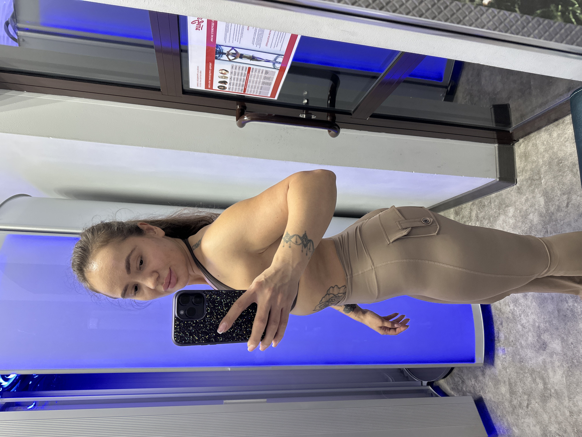 A woman with brown hair pulled back in a ponytail, wearing a dark sports bra and tan leggings, takes a mirror selfie. She is standing in a room with a tall, vertical tanning bed to her left, which emits a bright blue light. The floor is a light gray speckled tile. To her right is a glass door with a brown frame and a dark brown handle. A poster is affixed to the glass door, displaying images of people and text in a red and white color scheme. The woman's left arm is raised, holding a black phone with a sparkly case, and her right arm is bent at the elbow, resting on her hip. She has tattoos on her left arm and abdomen.