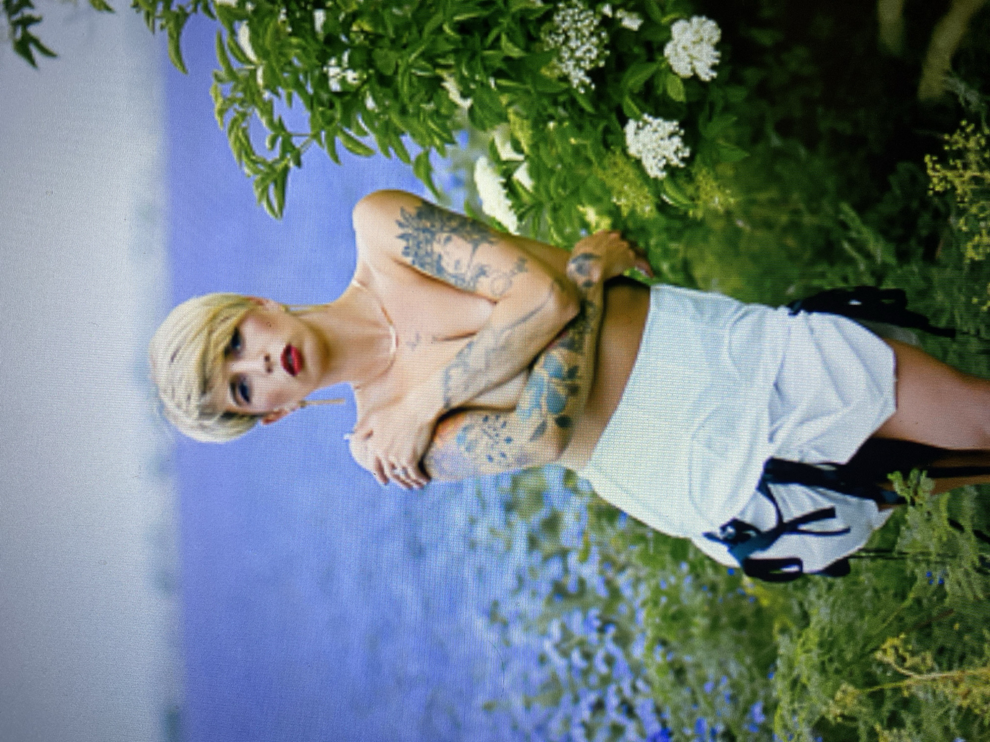 A blonde woman with short, choppy hair and red lipstick stands in a field of blue flowers and green foliage with a body of water in the background. She is wearing a white skirt with black ties. Her arms are crossed in front of her chest, and she has tattoos on her arms. The sky is overcast and appears to be a pale blue-grey. The overall scene is somewhat blurry, suggesting it may be a photograph taken with a phone or a low-resolution camera. The location is not specified, but the presence of a body of water, flowers, and foliage suggests an outdoor, natural setting, possibly a park or a rural area.