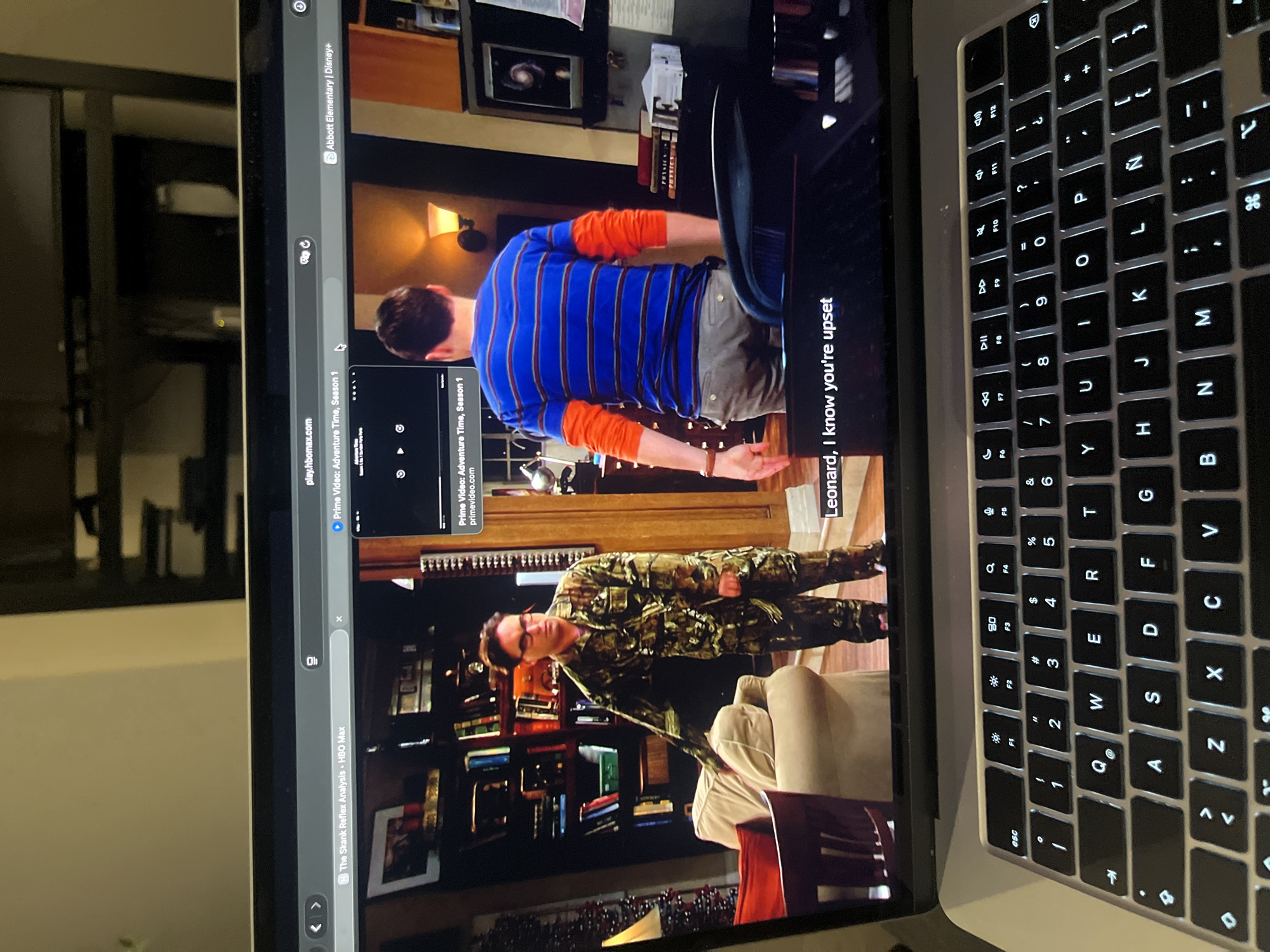 A laptop displays a scene from "The Big Bang Theory." The characters Sheldon and Leonard are visible. The browser bar shows tabs for "Abbott Elementary," "Prime Video: Adventure Time," and "The Skank Reflex Analysis- HBO Max." Part of the laptop keyboard is also visible. The location is not specified in the image.