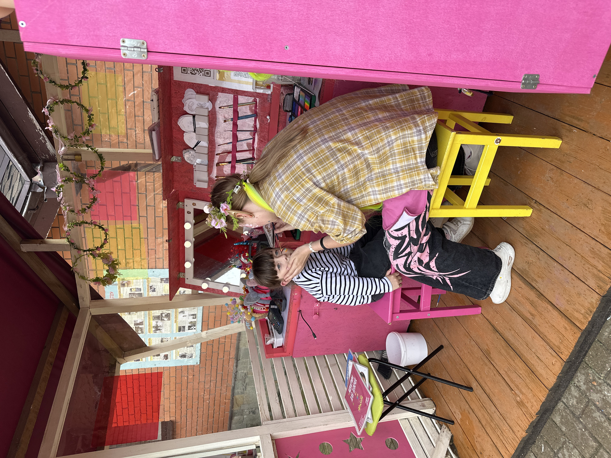 In Izhevsk, Russia, a young child with dark hair and blue eyes, wearing a black and white striped shirt, is having their face painted. They are sitting on a pink stool inside a colorful stall decorated with floral garlands and pink accents. A person, whose back is to the camera, is applying face paint to the child's cheek. This person has blonde hair tied in a ponytail, wears a yellow plaid shirt, and has a green accessory around their neck. The stall features a vanity with lights, makeup supplies, and brushes. The ground is made of wooden planks, and the stall is situated outdoors, possibly in a park or at an event, with a brick wall visible in the background.