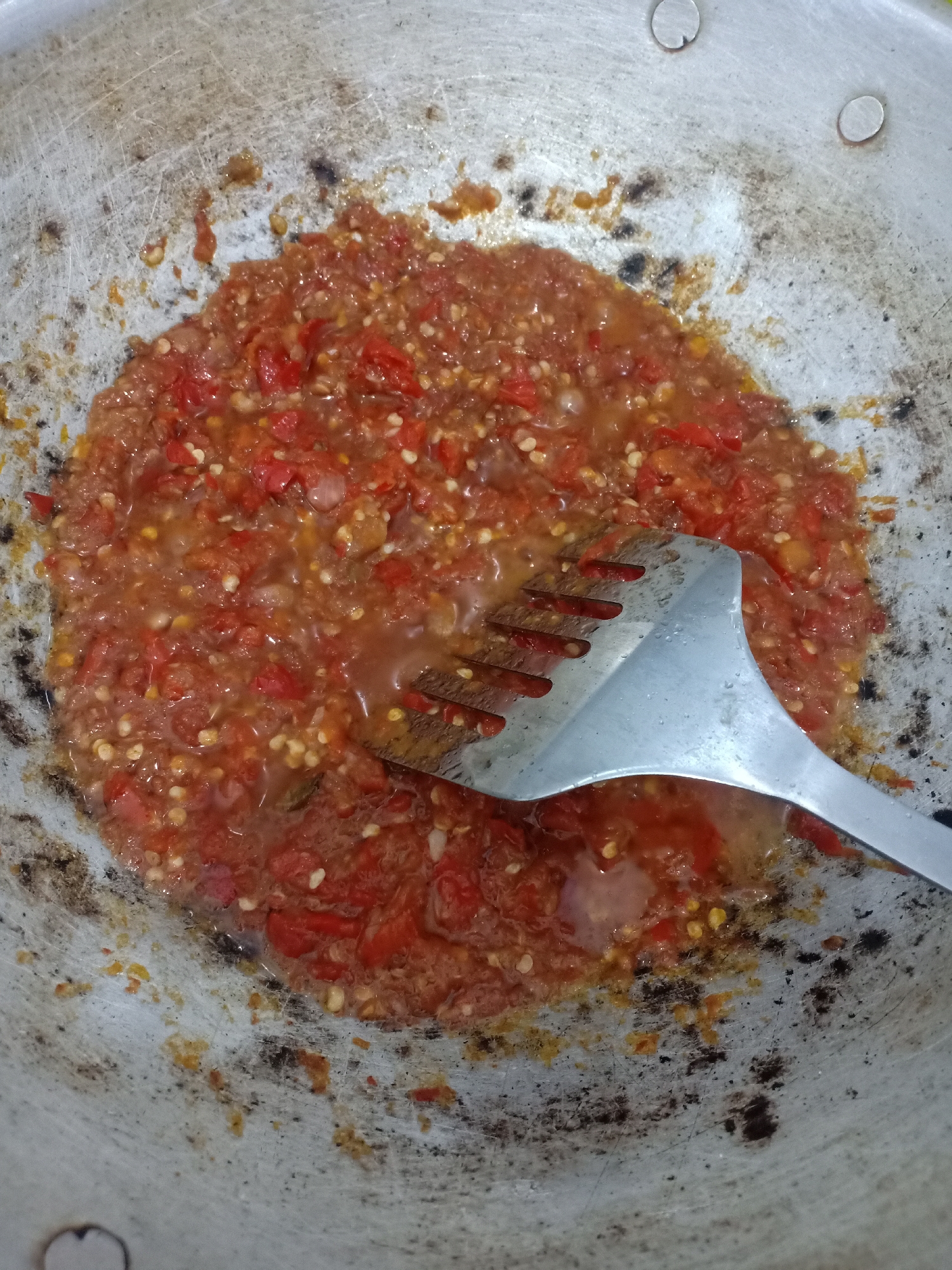 In a well-used, slightly scorched aluminum wok, a vibrant red chili paste is being cooked. The paste is chunky, with visible pieces of chili, seeds, and what appear to be finely chopped shallots or onions. A silver metal spatula with tines is placed in the center of the wok, partially submerged in the simmering mixture, suggesting an active cooking process. The wok's surface shows signs of wear, with some darkened, burnt-on residue and scratches. The surrounding environment is not visible. This scene is likely a common sight in kitchens across Semarang, Indonesia, where chili paste, known as sambal, is a fundamental part of many dishes.