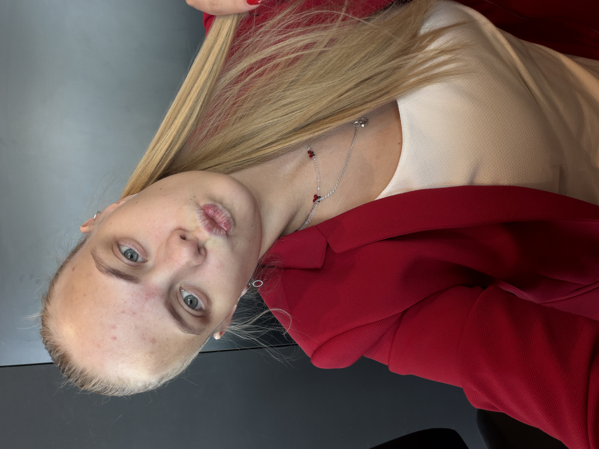A young woman with fair skin and blonde hair, is shown close-up. She has light eyes, fair eyebrows and slightly puckered lips. She is wearing a light-colored top and a red blazer. A silver necklace with red accents adorns her neck, and silver hoop earrings are visible. The background is a mix of grey and dark surfaces.
There is no indication of the location being Izhevsk, Russia.