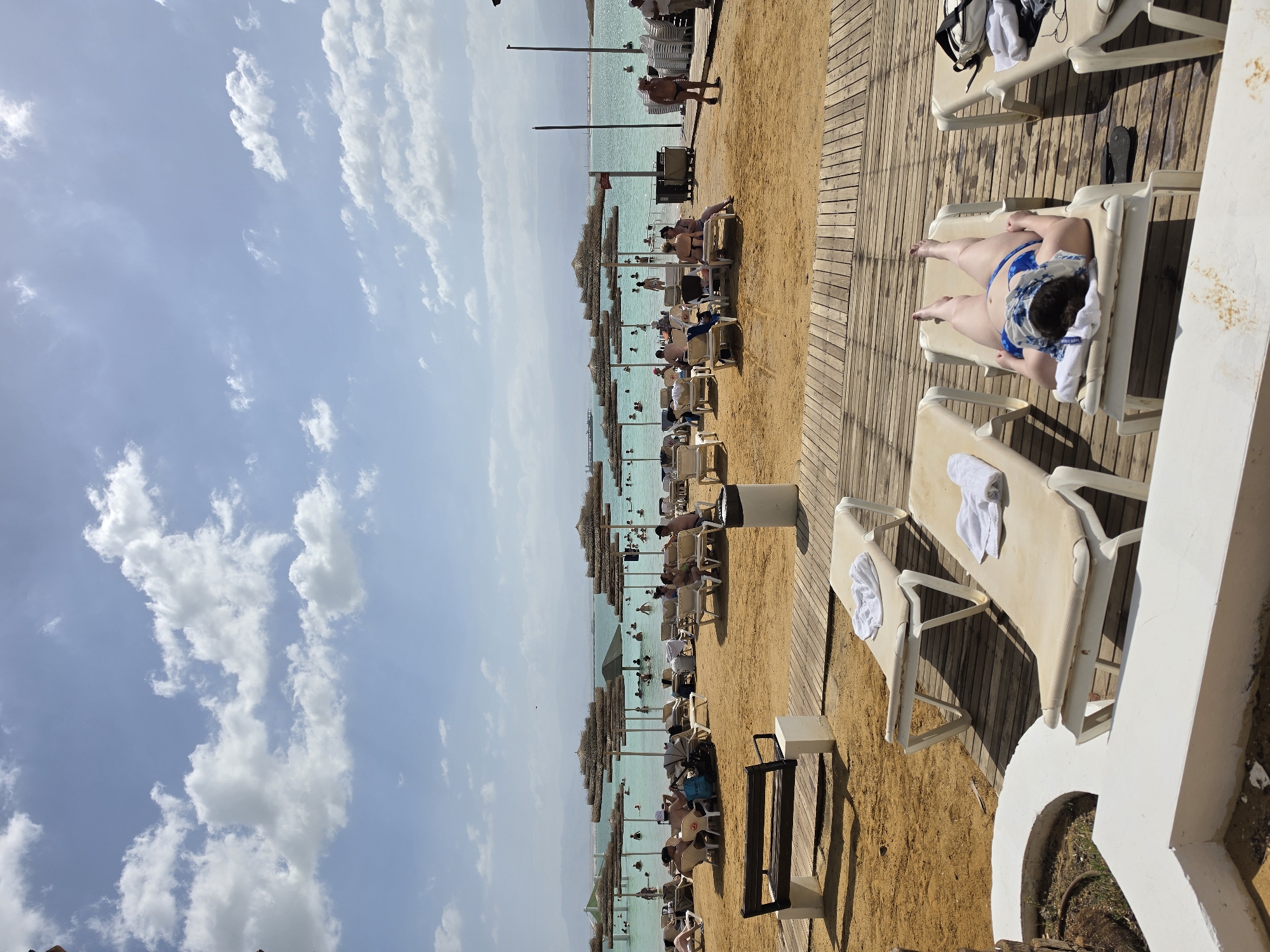The image depicts a beach scene in Tamar Regional Council, Israel. The foreground shows a wooden deck with several vacant lounge chairs and one occupied by a person wearing a blue swimsuit and a towel covering their head. Beyond the deck, a sandy beach is lined with numerous lounge chairs and sun umbrellas, most of which are occupied by people. The Dead Sea stretches out in the middle ground, with a hazy horizon in the distance. The sky is light blue with scattered white clouds.
