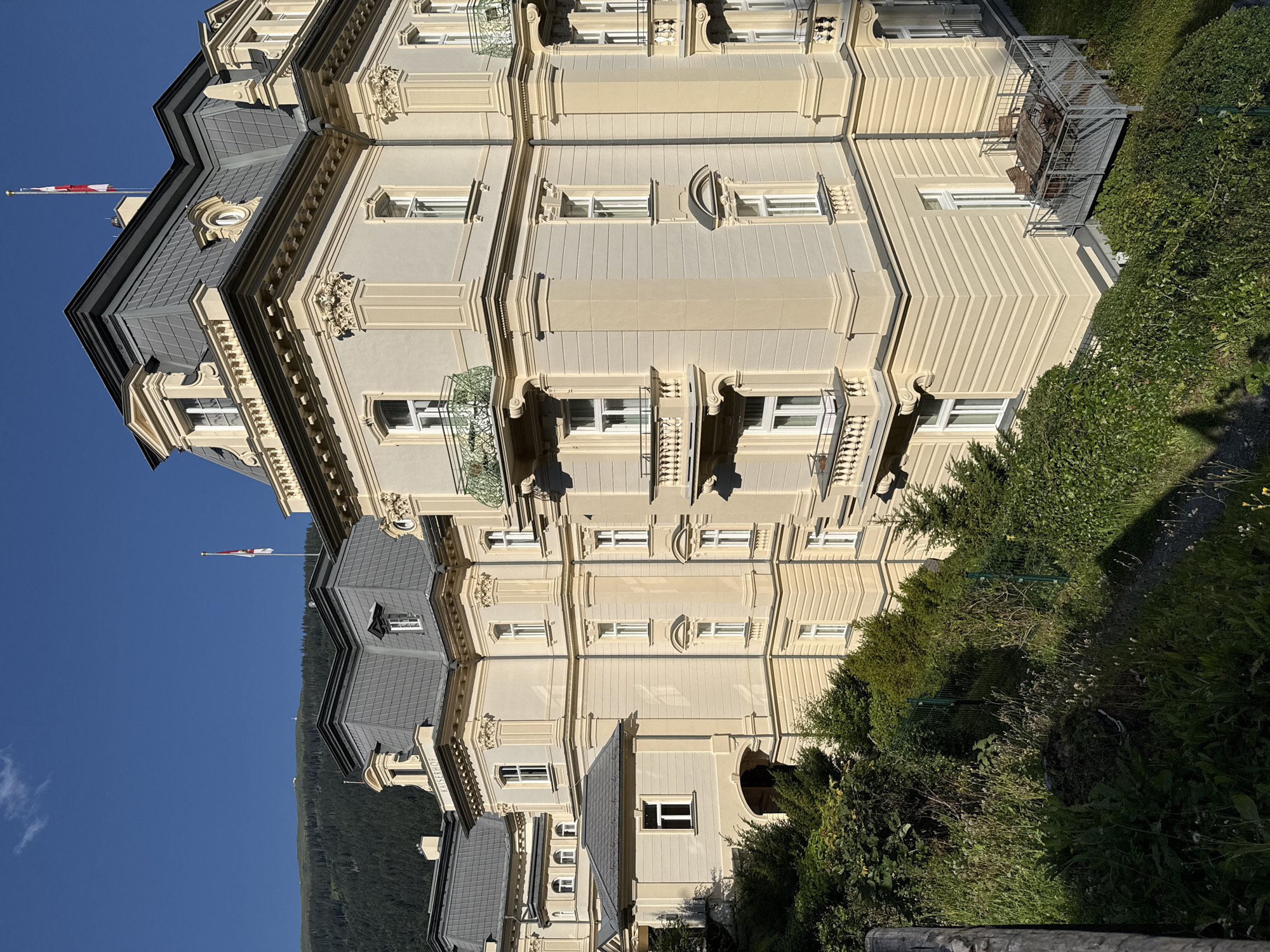The image shows the Kaiserhof Hotel in Bad Gastein, Austria, a grand Belle Époque building with a mansard roof and ornate detailing. The hotel is situated on a hillside, with lush green vegetation and trees in the foreground and a mountainous, forested landscape in the background under a clear blue sky. The hotel's facade is painted a pale yellow, with cream-colored trim and decorative elements. Several balconies with wrought-iron railings are visible, and one on the right side has outdoor furniture. The hotel has a prominent mansard roof with dormer windows and a flagpole flying a red and white flag. The architecture suggests a historic and elegant establishment.