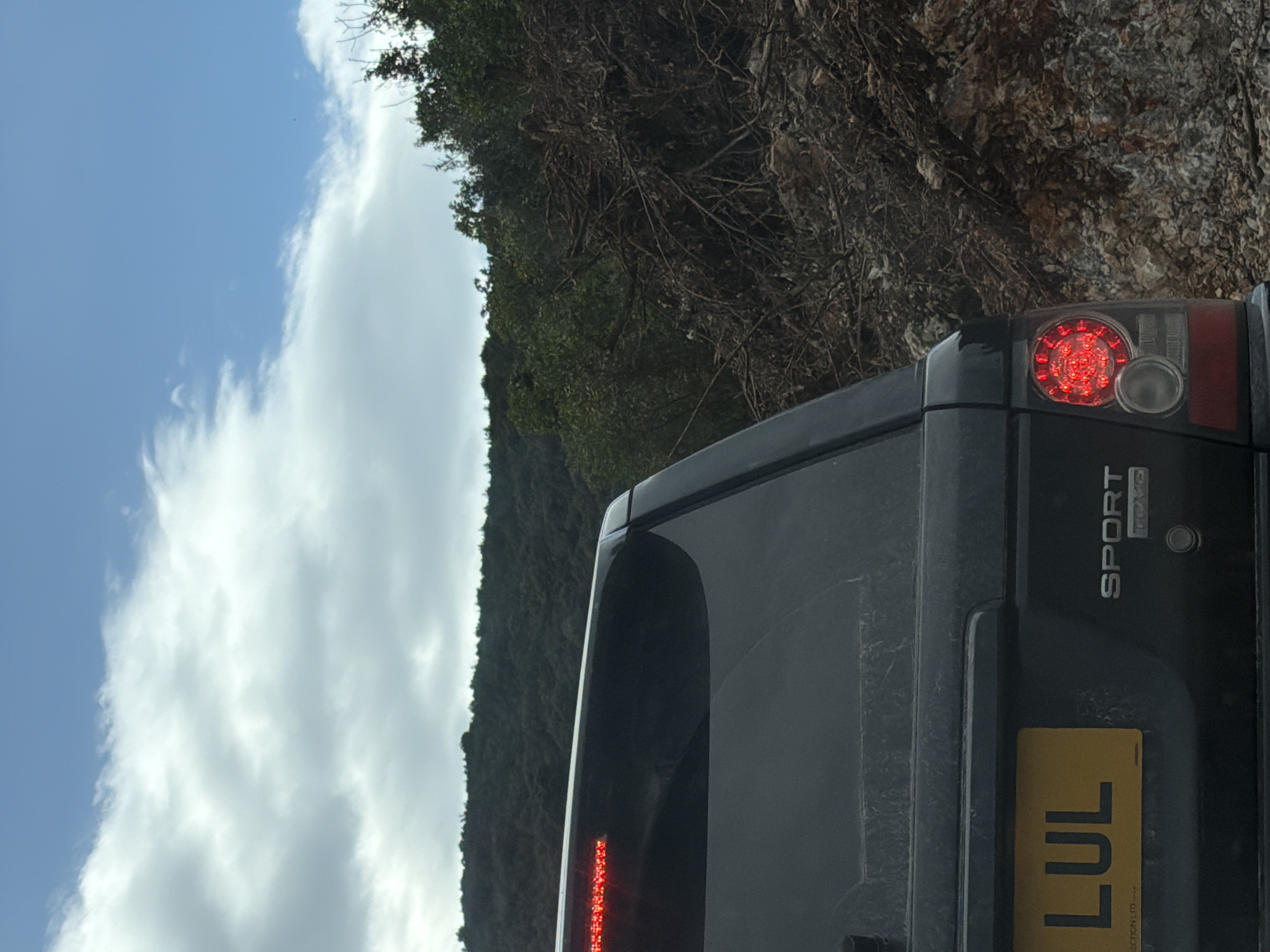 The image depicts the rear of a dark-colored Range Rover Sport in Radovići, Montenegro. The vehicle is positioned on a road adjacent to a steep, rocky hillside covered with dense vegetation. The sky is visible in the background, showing a mix of blue and white clouds. The vehicle's taillight is illuminated, and the license plate is partially visible, displaying the letters "LUL." The "Sport" model designation is visible on the right side of the vehicle.