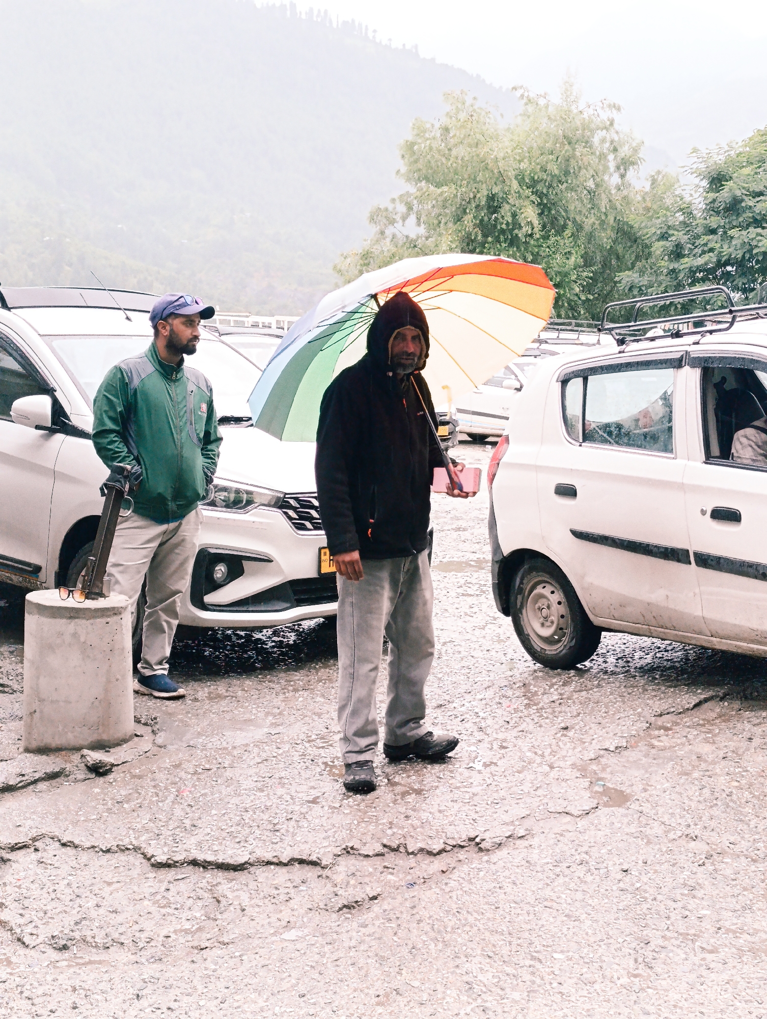 In Vashisht, India, two men stand next to two white cars on a wet, cracked asphalt surface. The man on the left, wearing a green jacket and a blue cap, stands with his hands in his pockets, holding a metal tool leaning against a concrete pillar. Sunglasses rest on the pillar. The man on the right, wearing a dark hooded jacket, holds a colorful umbrella over his head. The sky is overcast and a foggy mountain range is visible in the background. The scene suggests a slightly damp or rainy environment.