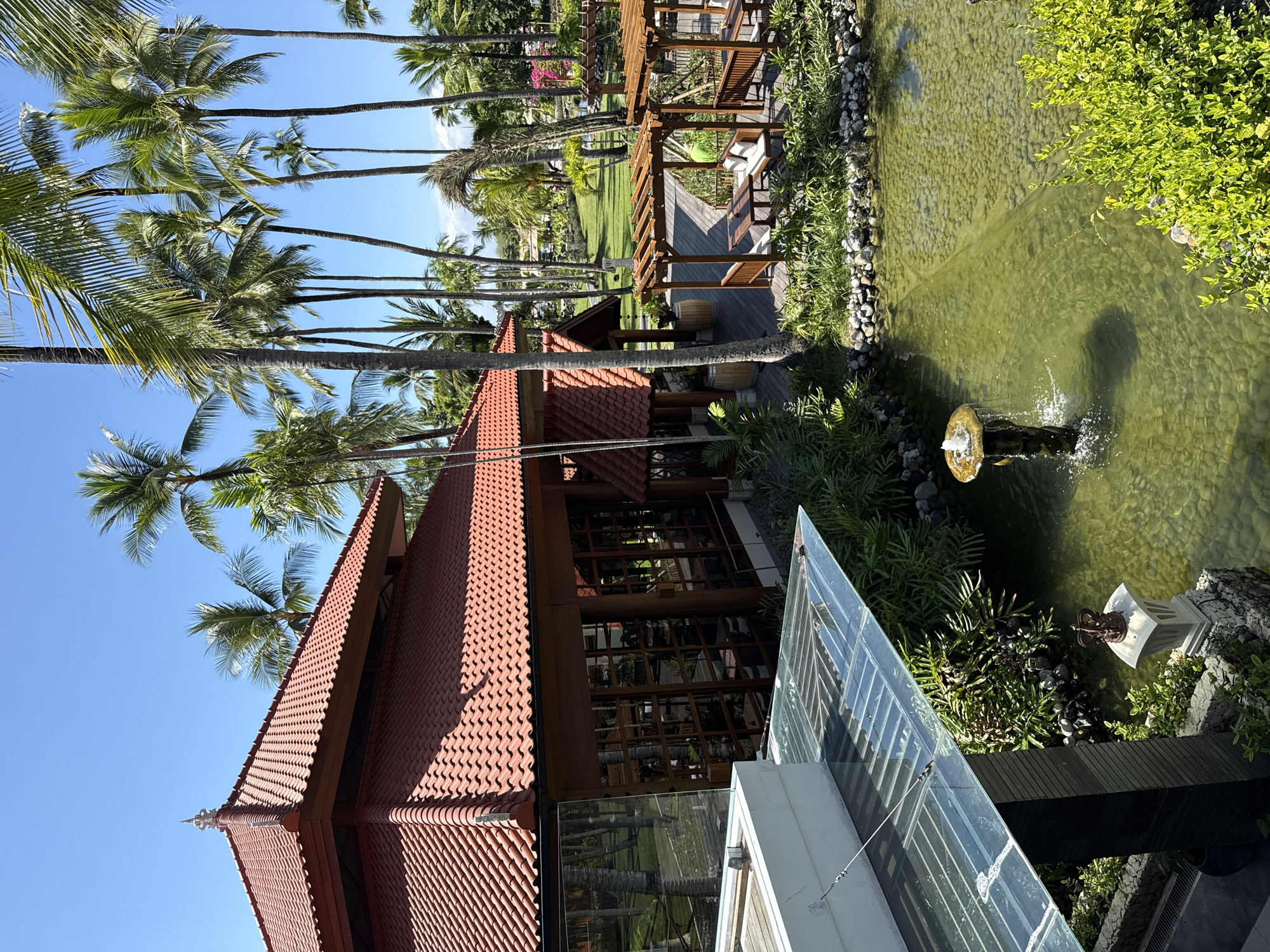 This vibrant image captures a serene tropical resort scene under a clear, bright blue sky, likely in Kuta Selatan, Indonesia. The prominent subject is a large building with a distinctive multi-tiered, red-tiled roof, characteristic of traditional Southeast Asian or Balinese architecture. Its facade features extensive wooden framework and large windows that reflect the bright surroundings.
To the right of the building, a long, narrow water feature resembling a pond or canal runs parallel. Its clear, greenish water reveals a bed of pebbles or small stones, and a small dark fountain ornament sprays water upwards near the foreground. The banks of the water feature are lush with green bushes and tropical plants.
Numerous tall palm trees with green fronds dominate the left side and background, creating a dense canopy against the bright sky. Beyond the main building and water feature, a well-maintained green lawn is visible, bordered by more palm trees and distant foliage. A wooden deck extends from the building, featuring a wooden pergola overhead and several wooden benches or seating arrangements. Two large wooden barrels are placed on the deck. Further back on the lawn, a lounge chair is visible under another wooden structure. A modern glass railing in the immediate foreground suggests a viewing platform or balcony.
The scene is bathed in bright sunlight, casting sharp shadows and indicating a clear, sunny day, likely late morning or early afternoon. No people are visible, contributing to an atmosphere of tranquil elegance and relaxation, typical of a luxurious tropical getaway. There is no visible text.